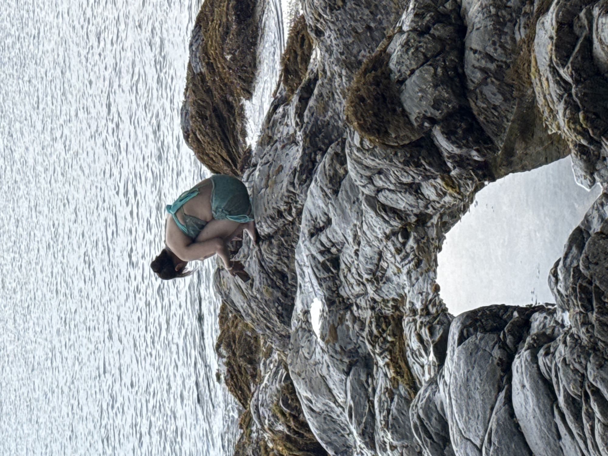 A woman is positioned on a rocky outcrop near the water in Đà Nẵng, Vietnam. She is wearing a teal-colored garment and is bending over, holding her legs close to her chest. The surrounding environment consists of jagged rocks and water, possibly the ocean. The perspective of the image is rotated, making the scene appear upside down. The rocks are heavily textured and mottled with patches of brown, likely indicating seaweed or other marine life.