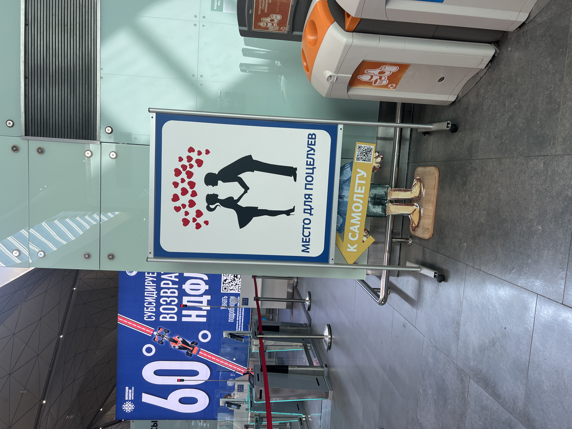 This is a photograph taken indoors, likely in an airport or transportation hub, given the presence of security gates and signage. The image is oriented vertically.
The main subject is a tall, white freestanding sign with a blue border. On the sign, a silhouette of a couple kissing is depicted, surrounded by a multitude of small red hearts. Above the couple, text in Russian reads "МЕСТО ДЛЯ ПОЦЕЛУЕВ" (Place for kisses). To the right of this text, on a yellow strip, is a QR code and the Russian words "К САМОЛЕТУ" (To the airplane).
In the background, a large blue banner displays the number "60%" prominently, along with Russian text that includes "СУБСИДИРУЕ" (subsidized) and "ВОЗВРА" (return) and "НДФЛ" (NDFL, likely referring to Personal Income Tax). This banner appears to be advertising a financial incentive or program.
To the right of the kissing couple sign, there is a recycling bin with orange accents, featuring graphics of bottles and cans. In the foreground, red velvet ropes guide people through security checkpoints. The floor is tiled with large, gray square tiles.
The overall scene suggests a public space, possibly an airport, where travelers are preparing to depart. The "Place for kisses" sign adds a touch of romance and humor to the functional environment, while the large banner advertises a significant financial benefit. The lighting appears to be artificial, consistent with an indoor setting.