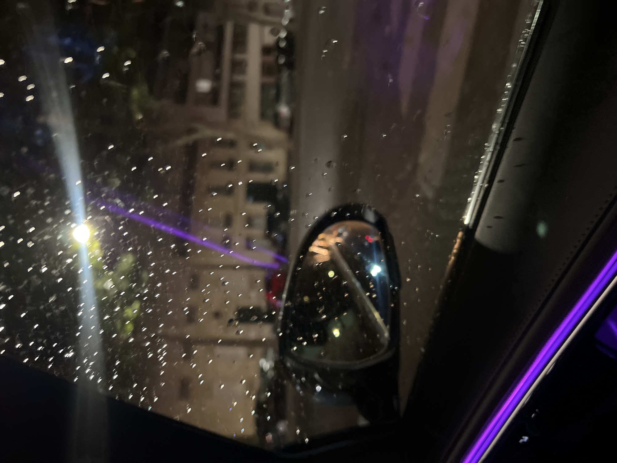 The image depicts a view from inside a vehicle at night, looking out the passenger window. Raindrops are visible on the glass, creating small, distorted points of light from the surrounding environment. A side-view mirror is prominent in the foreground, reflecting city lights and some street activity. Beyond the mirror, the background shows a street scene with buildings, parked cars, and a streetlamp casting a bright, diffused light. The streetlamp also appears to have a purple light source reflecting off it, or a similar light source in the background. There is a noticeable purple ambient light emanating from within the car, illuminating the door panel in the lower portion of the image. The scene suggests a city environment, possibly Paris, France, given the architectural style of the buildings. No people are clearly identifiable in the image due to the low light conditions and the rain distorting the view.