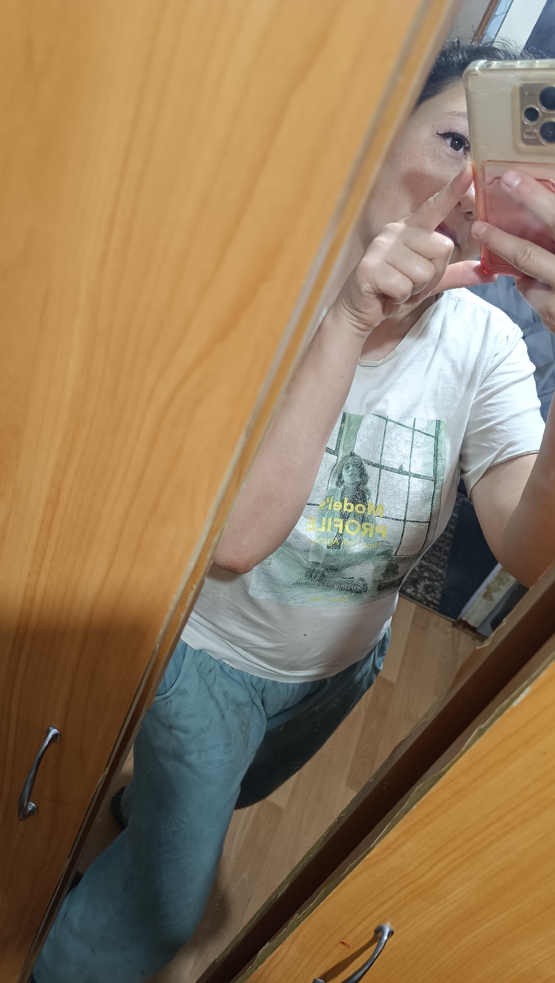 The image shows a reflection of a person in a mirror, taken from a high angle. The person is wearing a white t-shirt with a graphic print and light blue pants. They are holding a smartphone in their right hand, pointed towards the mirror, and touching the mirror with their left index finger. The reflection shows the person's face with eye makeup, as well as their torso and legs. In the background, a wooden dresser or cabinet is visible, with a drawer pull on the left side. The environment appears to be indoors, possibly a bedroom or dressing area. The image does not provide any information to indicate the location is Karsha, Russia.
