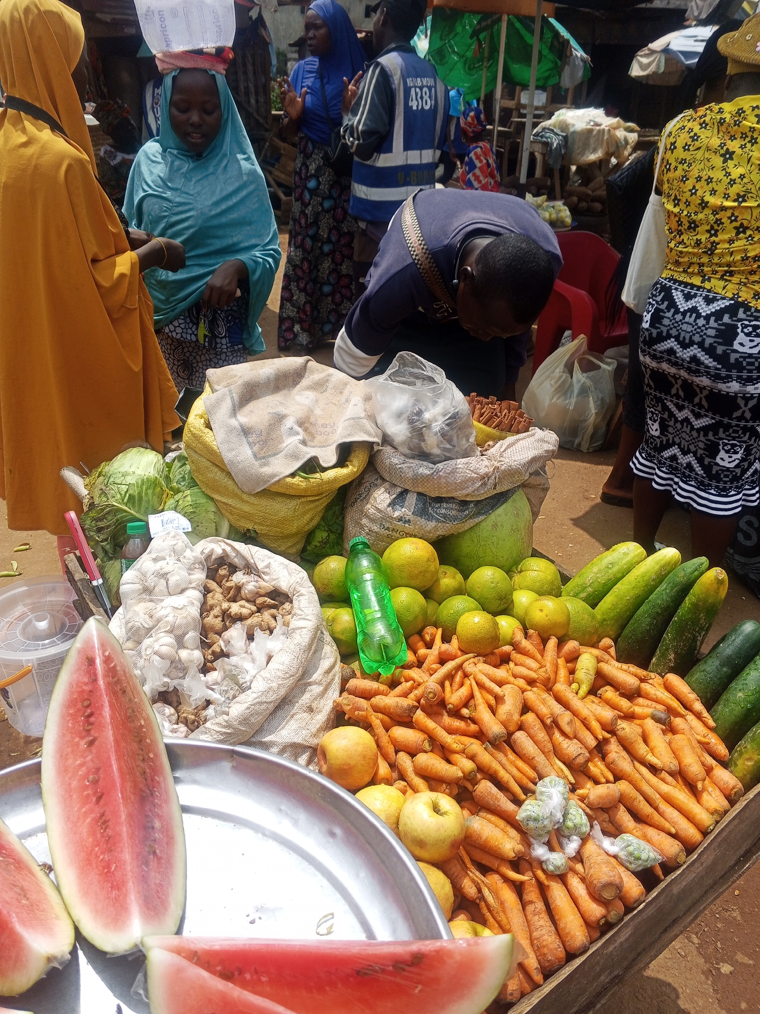 The image depicts a vibrant market scene in the city of Kabba, Nigeria. In the foreground, sliced watermelons are arranged on a silver platter. Behind the watermelons, a large display of fresh produce is laid out. This includes piles of bright orange carrots, several green cucumbers, and a mound of round, yellow citrus fruits, likely oranges. A bag filled with what appears to be garlic or ginger is also visible. In the background, various individuals are present. One person, wearing a yellow head covering and a matching robe, stands to the left. Another individual, in a blue head covering, is conversing with the person in yellow. Further back, someone in a dark blue top with a white and brown striped strap across their chest is bending over, possibly examining the goods. Other market stalls and shoppers are visible in the distance, creating a bustling atmosphere typical of a Nigerian marketplace. The overall setting appears to be an outdoor market, with a dirt ground and various structures suggesting temporary stalls.