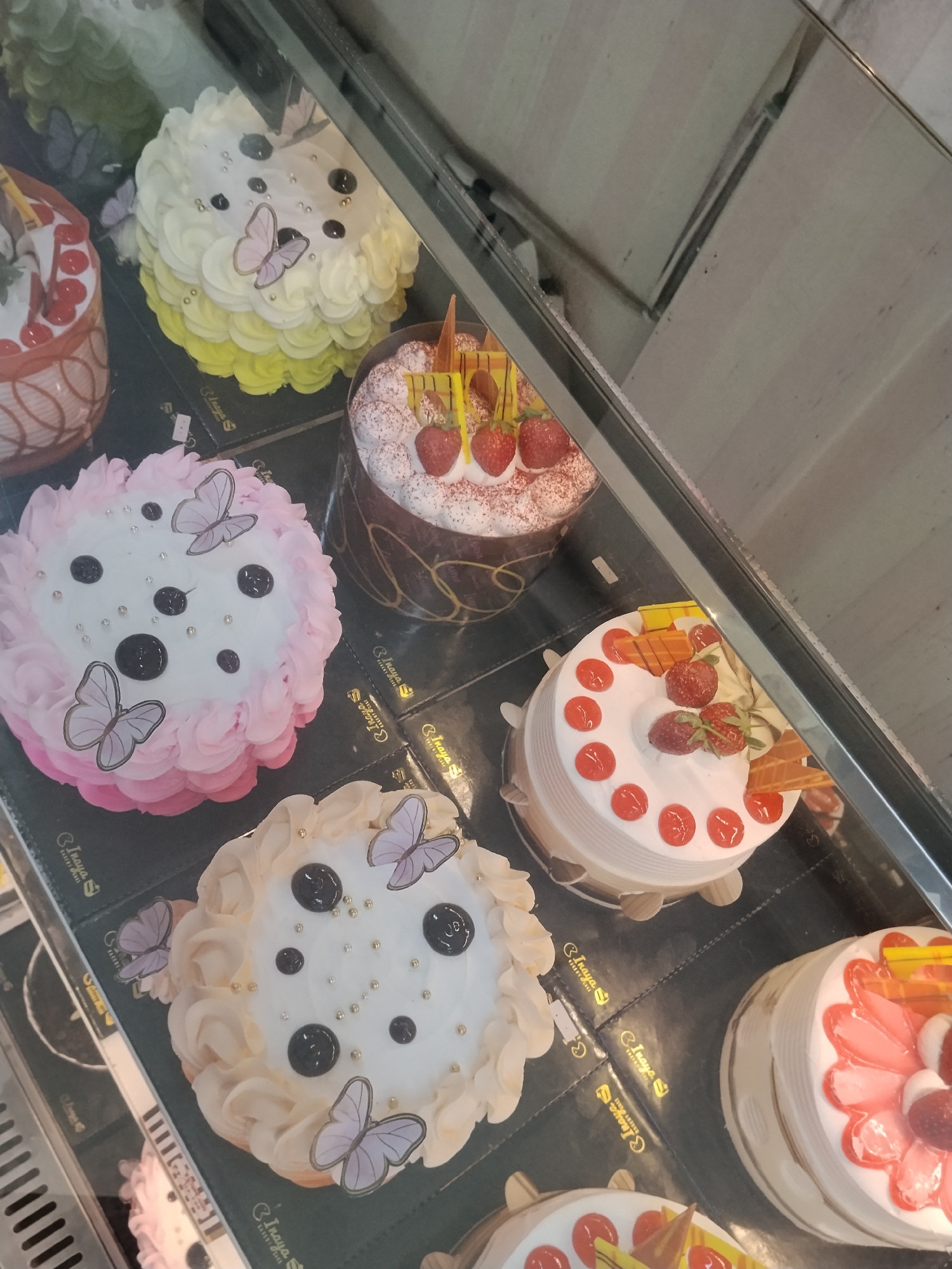 The image displays a selection of cakes arranged on shelves within a bakery display case. The cakes vary in decoration and color, with some featuring floral frosting, fruit toppings such as strawberries, and decorative butterflies. The overall impression is of a commercial bakery setting. The exact location within Ciruas, Indonesia, is not discernible from the image itself, as it lacks any specific geographical markers. The scene is static, with no people present and no actions occurring within the frame. The environment appears to be an indoor retail space.