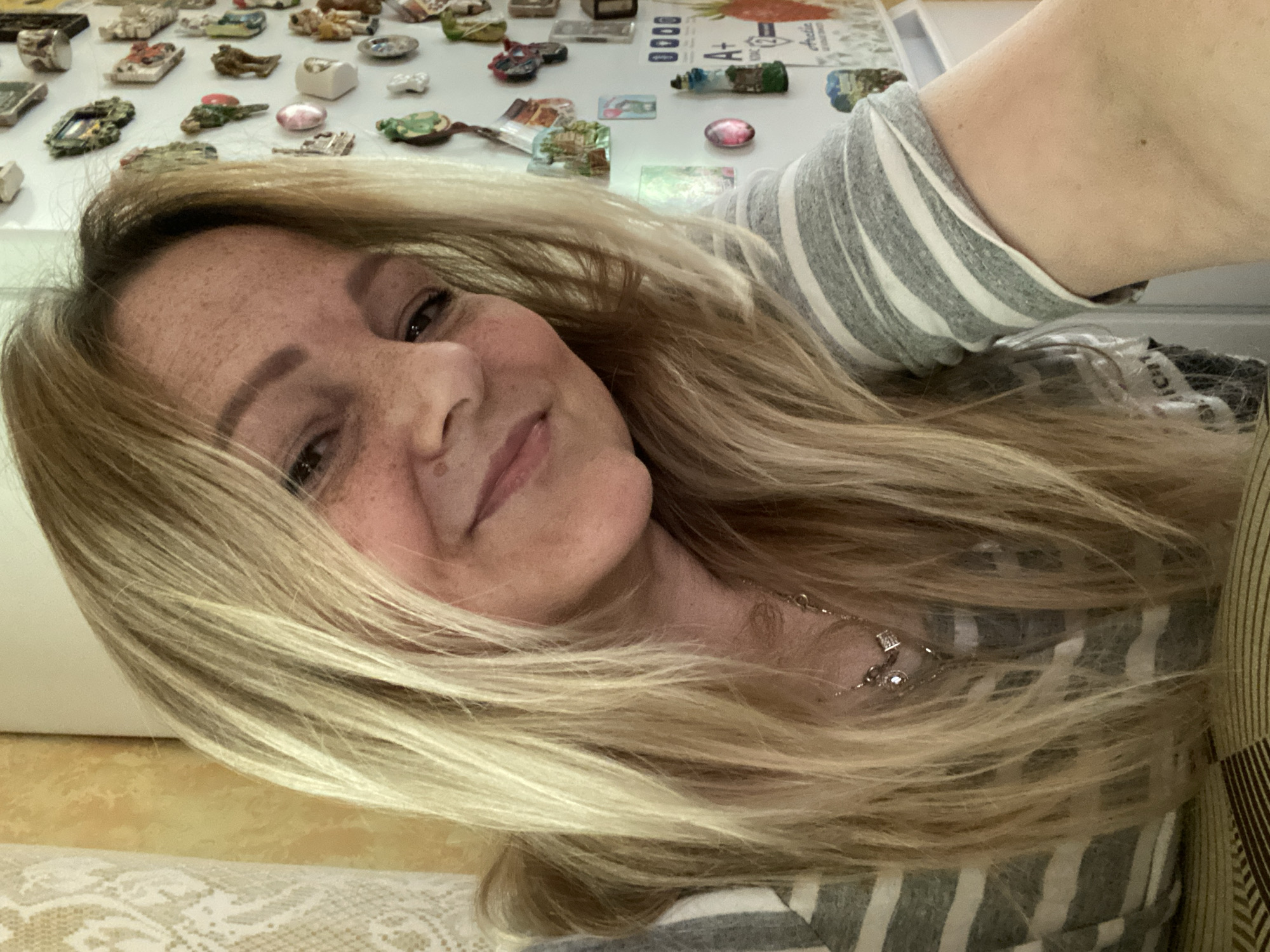 The image shows a close-up selfie of a woman with blonde hair and freckles, taken indoors. She is wearing a gray and white striped shirt and a necklace. The background shows a wall covered with numerous colorful magnets, suggesting the location is likely a kitchen or a similar domestic setting. There is also a glimpse of a patterned surface in the foreground, possibly a table. The scene does not provide any visual information to indicate the specific location within the city of Luh, Ukraine.