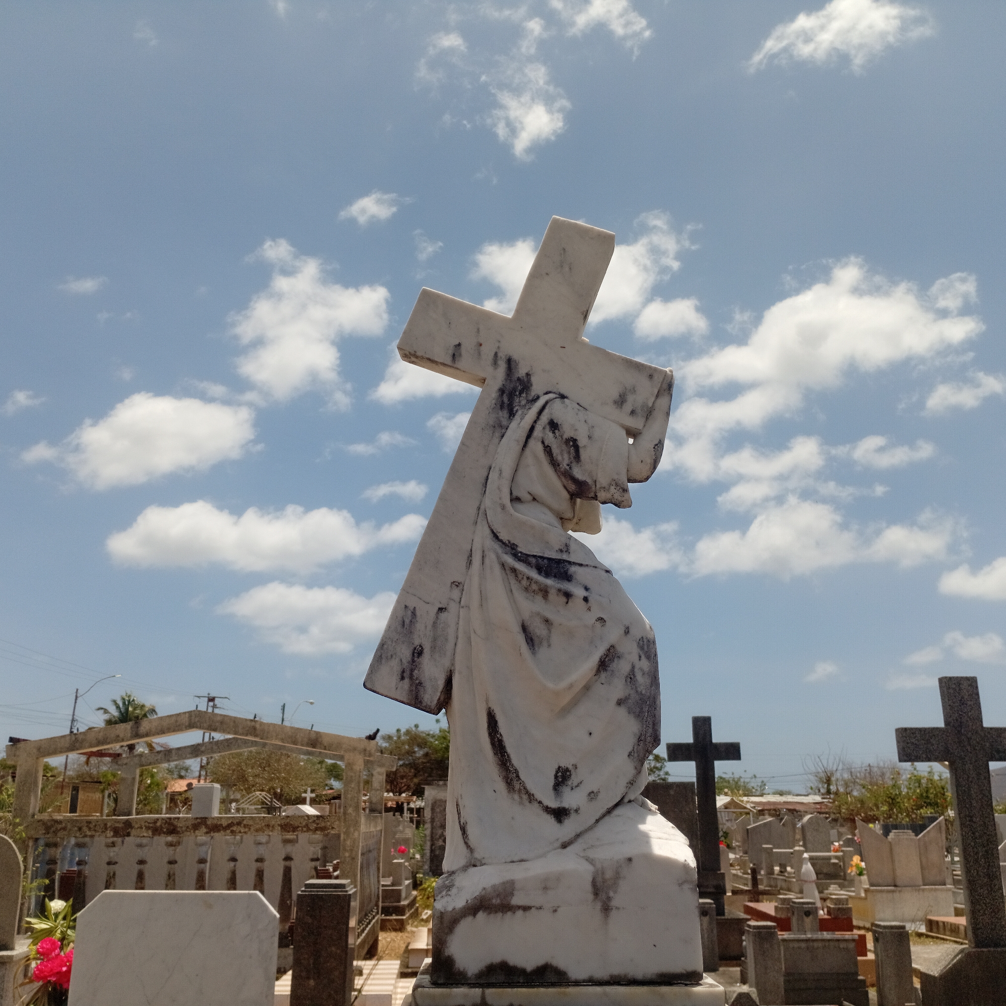 A marble statue of Jesus carrying the cross stands in a cemetery under a cloudy sky. The statue is positioned in the foreground, with the cross resting on Jesus's shoulder. The cemetery stretches into the background with various tombstones and crosses visible. The location appears to be in a city due to the presence of buildings and power lines in the distance. The specific city is Unknown, Unknown.