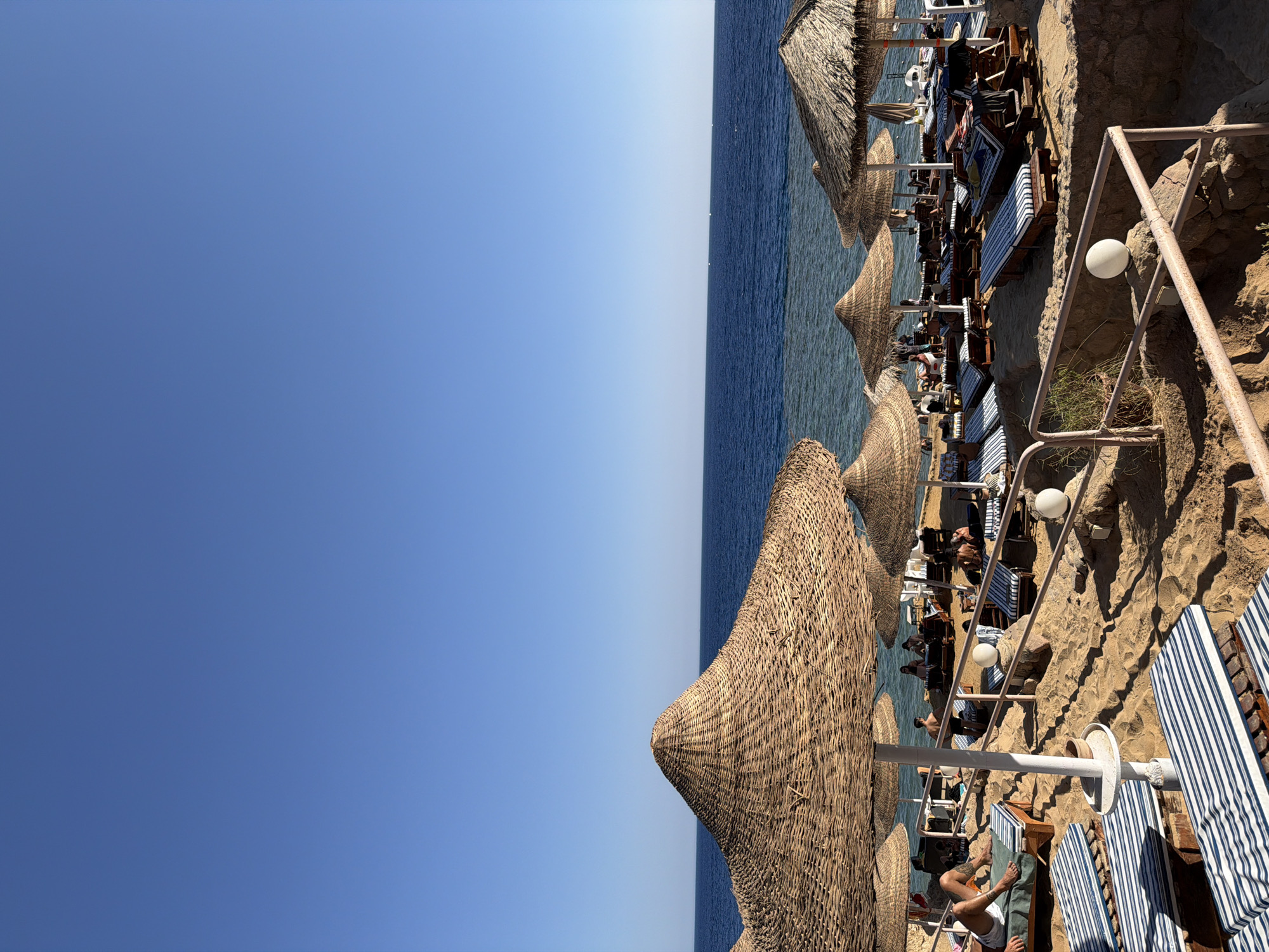 The image depicts a crowded beach in Sharm El Sheikh, Egypt, on a clear, sunny day. The foreground is dominated by a collection of thatched-roof umbrellas providing shade over numerous lounge chairs arranged on the sandy beach. Several people are seen relaxing on these chairs, some appearing to sunbathe. Further back, closer to the water, more umbrellas and lounge chairs are visible, as are groups of people. The sea is a deep blue, with the horizon clearly demarcated against a bright blue sky. The water appears calm, with some areas of shallow, clear water near the shore where the seabed is visible. The overall scene conveys a typical resort atmosphere, with recreational activities and leisure evident.