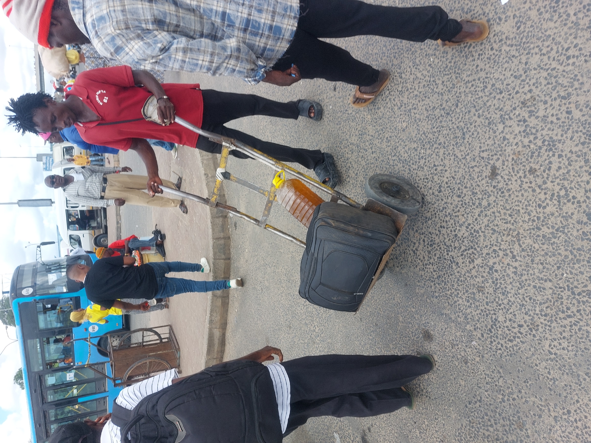 The image depicts a street scene in Dar es-Salaam, Tanzania. A man wearing a red shirt pulls a two-wheeled cart containing a suitcase and a large plastic bottle filled with an amber liquid. Other individuals are present, including a man in a blue plaid shirt, and others standing near a blue bus. The environment appears to be a busy street or transportation hub with pedestrian traffic. The road surface is paved, and buildings are visible in the background.