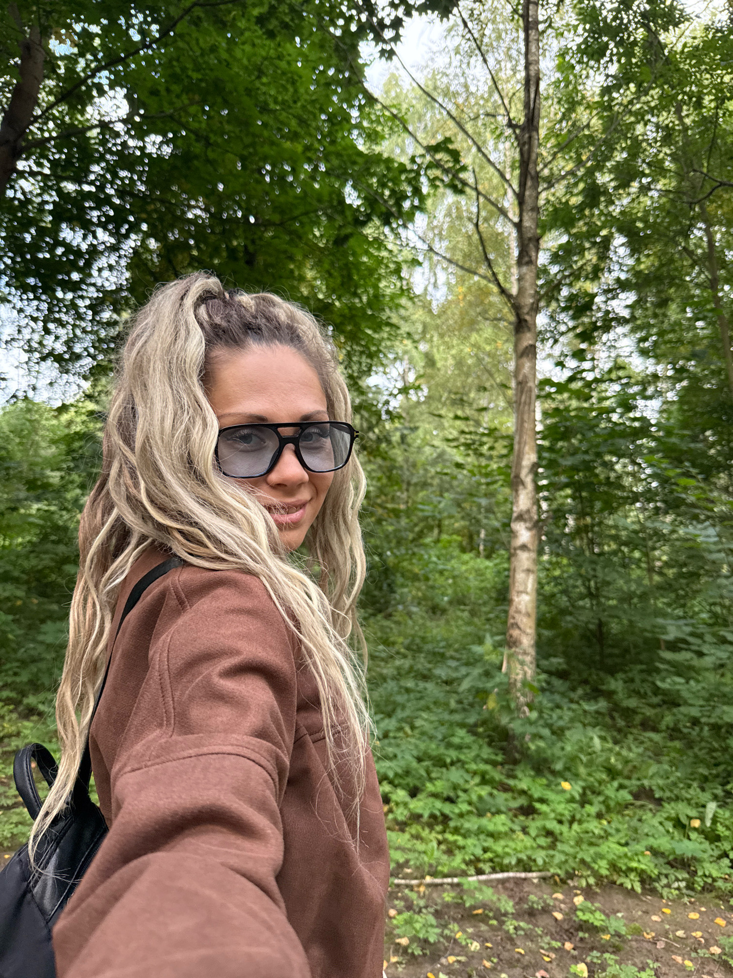 In Rusanovo, Russia, a blonde woman with long, wavy hair tied in a ponytail and wearing large dark-rimmed glasses and a brown hooded sweatshirt is captured in a self-portrait. She is standing in a lush, green forest setting. The background is filled with dense foliage, trees, and dappled sunlight filtering through the leaves, suggesting an outdoor, natural environment. A black backpack is visible on her left shoulder. The woman is looking towards the camera and smiling slightly, her expression relaxed and natural. The overall scene conveys a sense of casual outdoor activity.