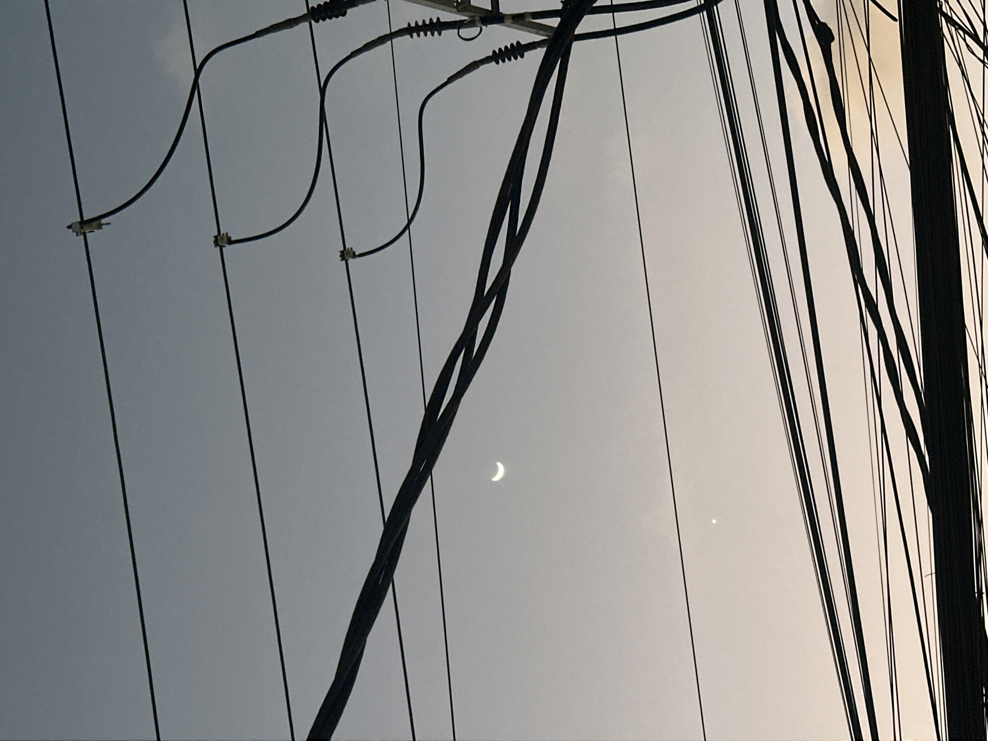 This image depicts a twilight sky over North Kuta, Indonesia, with a crescent moon and a bright star visible. The sky is a gradient of muted gray and pale yellow. A dense network of black electrical wires dominates the foreground and midground, crisscrossing the frame in multiple layers. Some wires are supported by utility poles, while others appear to be suspended independently. The wires are in focus, creating a stark contrast with the soft, diffused light of the sky. There are no people or any discernable actions or interactions. The scene is entirely focused on the celestial bodies and the infrastructure of overhead wiring.