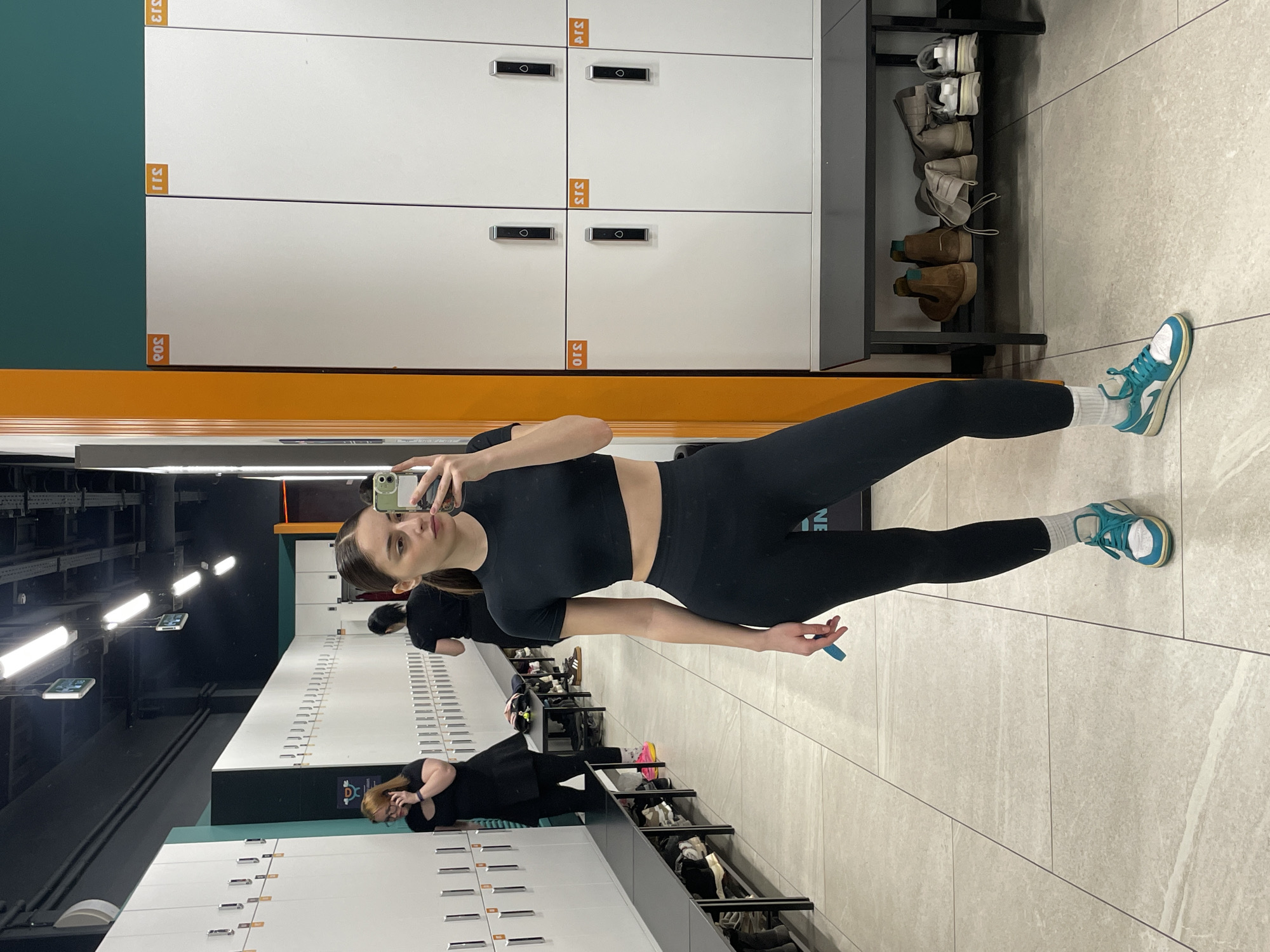 The image displays a modern locker room within a fitness facility, consistent with a location in Moscow, Russia. In the foreground, a young adult female with dark hair pulled back, wearing a black athletic crop top, black leggings, white socks, and white and teal athletic shoes, stands facing a mirror. She holds a smartphone with a green case in her right hand, taking a selfie, and a small blue object in her left hand.
The room features an aisle flanked by rows of white lockers, stacked two high, each equipped with a black electronic lock and an orange square sticker displaying three-digit Arabic numerals (e.g., '301' through '304' on the left, and mirrored '305', '311', '312', '313' on the right). Dark-colored benches run along the base of the lockers, with various footwear including sneakers, boots, and other shoes stored underneath.
In the background, on the left side of the aisle, a second female with reddish-brown hair, dressed in a black top and leggings, is observed speaking on a dark-colored smartphone. Her left foot reveals a bright pink shoe. Further down the aisle, a third female, also in black attire, is partially visible from behind.
The flooring consists of large, light-colored rectangular tiles. The ceiling is dark and industrial, featuring exposed conduits and multiple rectangular fluorescent light fixtures, along with some small hanging display screens. Accent elements include a dark teal wall section and a vertical orange panel. The lighting is bright and even throughout the area.