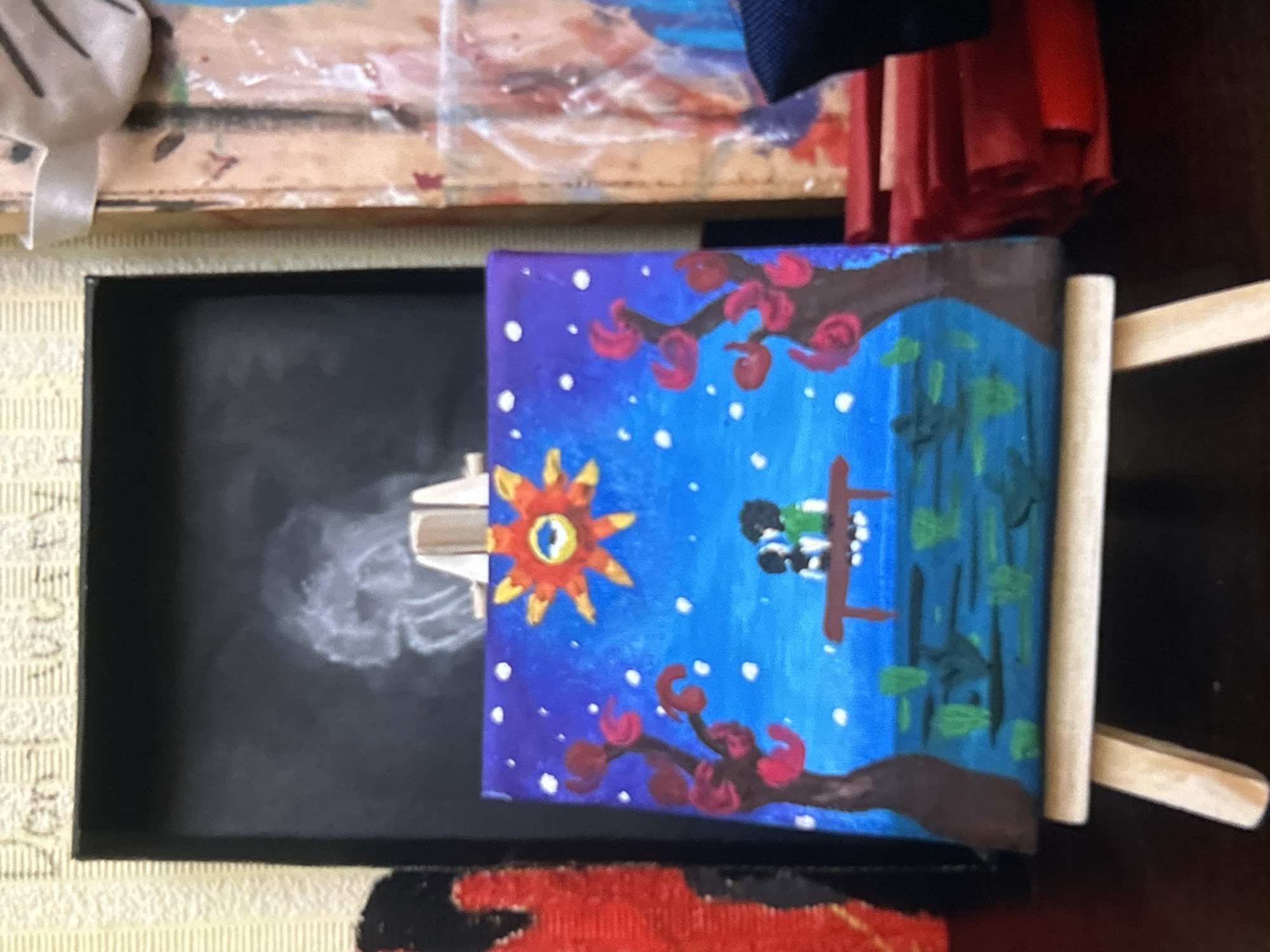 A small, amateur painting on a mini-canvas is propped up within a black box. The painting depicts a surreal scene: a stylized orange sun with a single eye sits against a blue, star-filled sky. Below, two figures are on a swing hanging from a tree with red blossoms. A dark, shadowy area possibly meant to be water is to the right. The box containing the painting is situated on a patterned surface. Other objects are vaguely visible in the background, including a wooden tray and some pieces of red fabric. The exact location of Tbilisi, Georgia is not identifiable from the image.