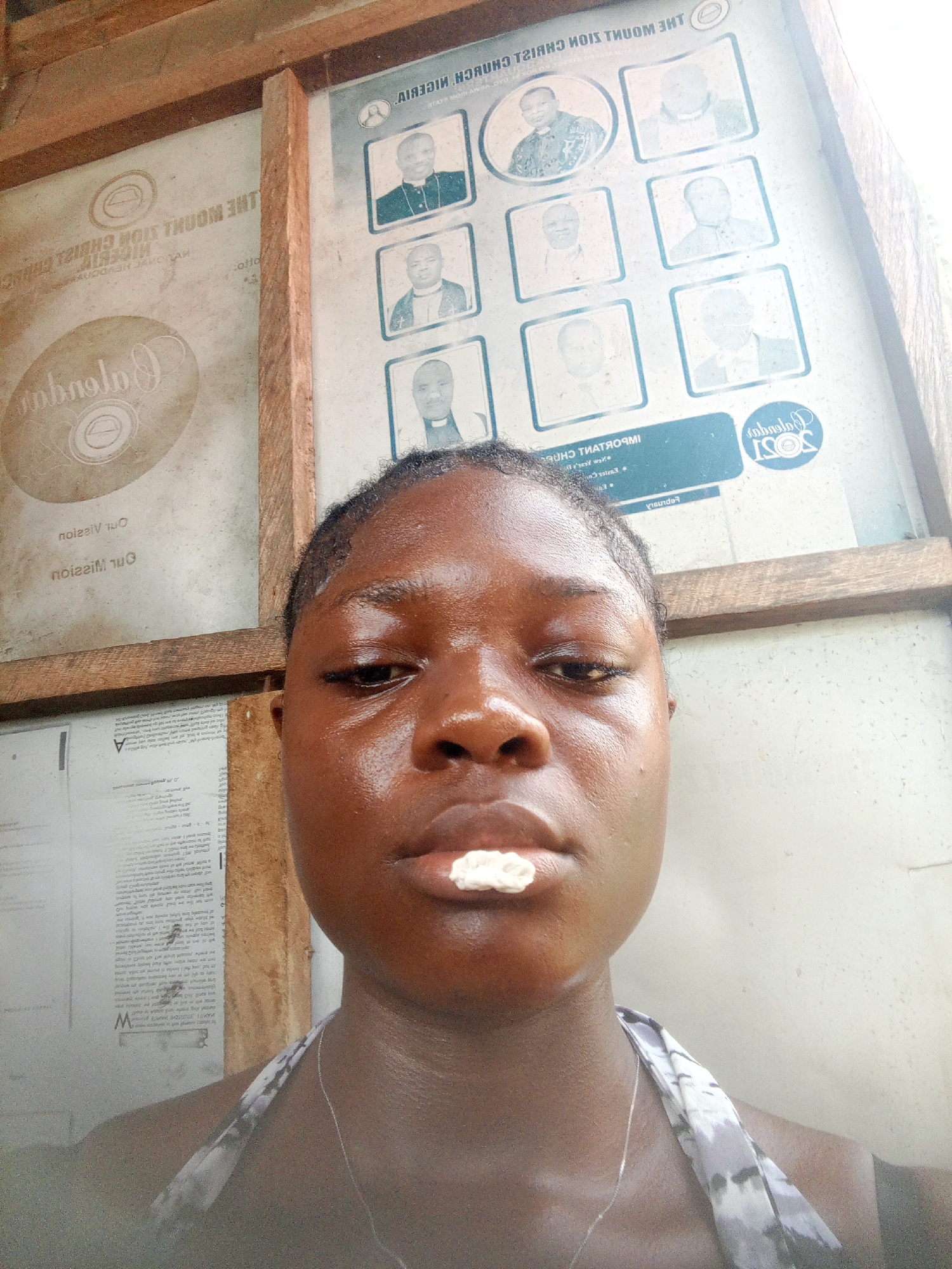 A close-up selfie shows a dark-skinned young person with a piece of white material (possibly gum or a medicinal substance) between their upper lip and teeth. They are wearing a halter top and a thin necklace. Behind the person, there are two wall-mounted signs within a wooden frame. The sign on the right features nine images of men, likely clergy members, and the text "THE MOUNT ZION CHURCH NIGERIA". To the left is another sign with an emblem and reversed text, including the word "Obanolo" and phrases "Our Mission" and "Our Vision" underneath. A third sign, with text and images, is visible underneath the sign on the left. The setting appears to be indoors, perhaps a church building. No specific details directly indicate the image was taken in Mbribit, Nigeria, although the church name suggests a Nigerian location.