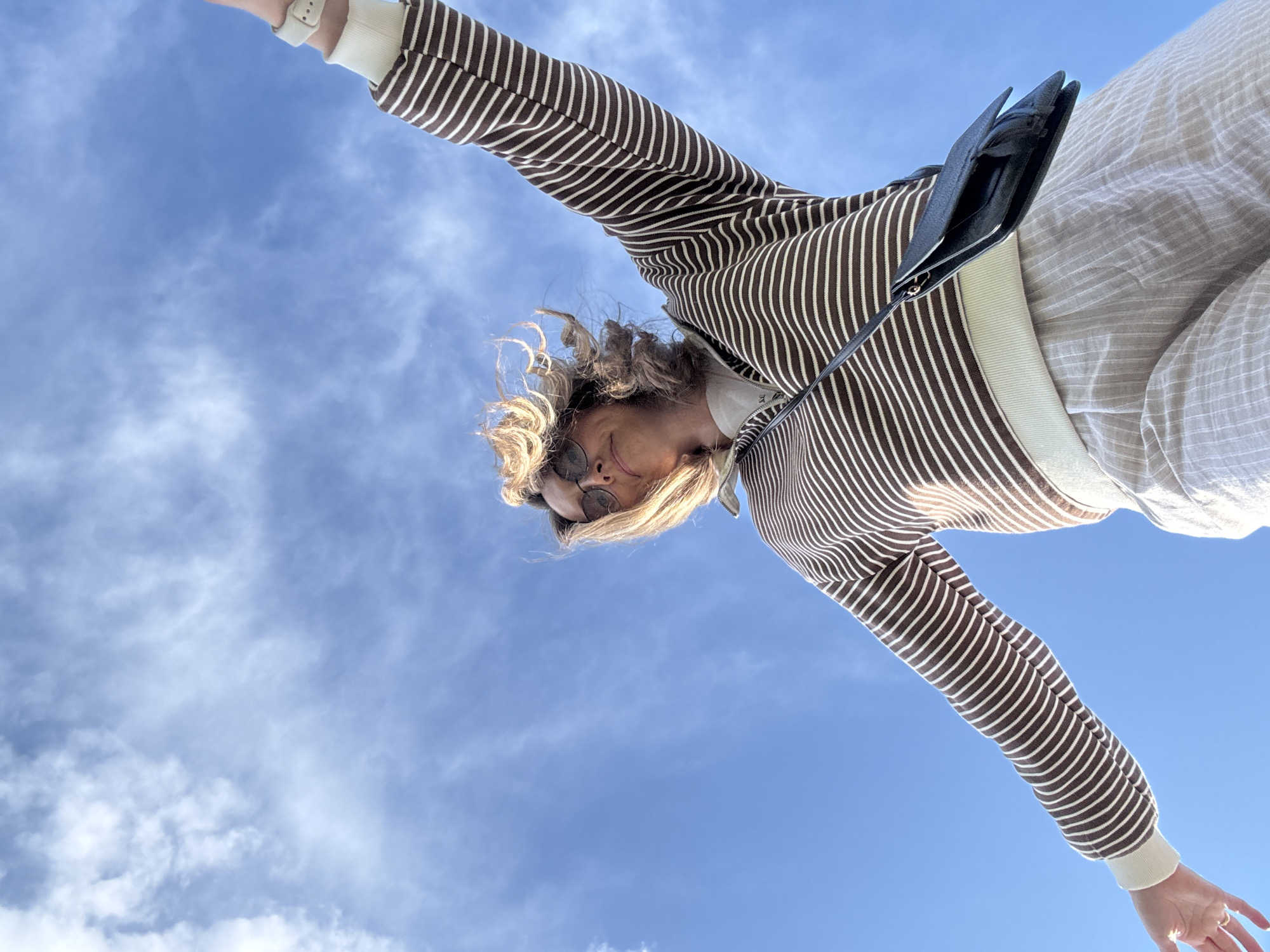 The image is taken from a low angle looking upwards, featuring a person against a blue sky with scattered clouds. The person is wearing a brown and white striped sweater, beige trousers, and sunglasses. They have curly blonde hair and a black bag hanging off their side. Their arms are outstretched. The location is not discernible, and it's not possible to determine if this image was taken in Votkinsk, Russia, based on the visual information.