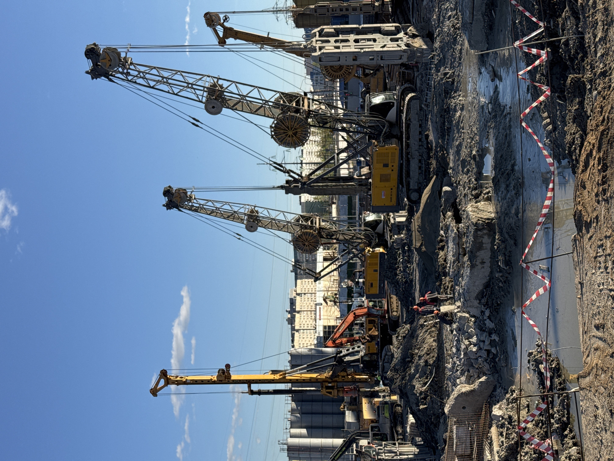 The image shows a construction site in Saint Petersburg, Russia, with several large crane-like machines dominating the scene. These machines feature long, latticed booms extending upwards against a clear blue sky with scattered clouds. In the middle ground, construction workers wearing helmets are visible near an orange excavator and other machinery, amidst a landscape of dirt, rocks, and construction debris. A newly-built gray structure runs parallel with a red and white barricade. In the background, a cluster of cylindrical industrial silos and low-rise buildings are visible, confirming the urban setting.