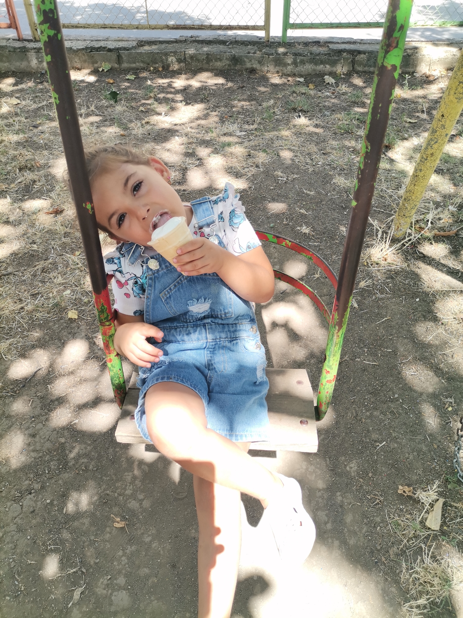 A young girl with blonde hair is sitting on a swing, eating an ice cream cone. She is wearing denim overalls over a short-sleeved shirt with a print. The swing has a wooden seat and metal supports, one of which is green and red, and the other is yellow. The ground beneath the swing is covered with dirt and dry grass. In the background, there is a metal fence and a concrete curb. The image appears to have been taken outdoors in a park or playground. The sun is casting shadows on the ground. The location is not identifiable from the image itself.