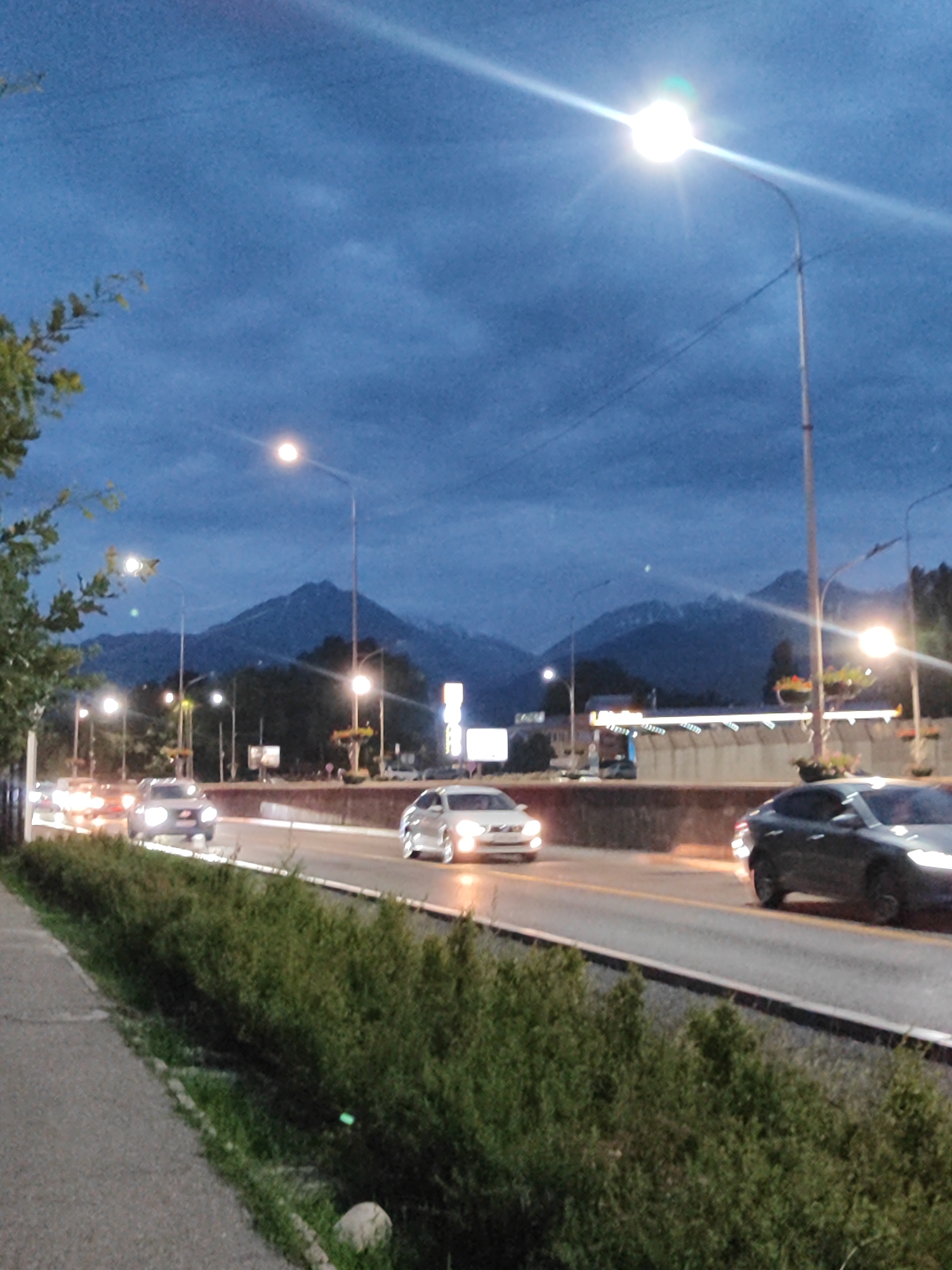 The image depicts a highway scene at dusk in Almaty, Kazakhstan. The sky is a deep blue with scattered clouds, indicating a transition from daylight to night. A bright streetlight illuminates the foreground with a lens flare. The highway is lined with trees and bushes, and several cars are visible on the road, their headlights and taillights glowing. In the background, mountains are silhouetted against the twilight sky, suggesting a mountainous landscape surrounding the city. The overall mood is peaceful and serene, with the artificial lights of the highway contrasting with the natural beauty of the mountains. There is no visible text in the image.