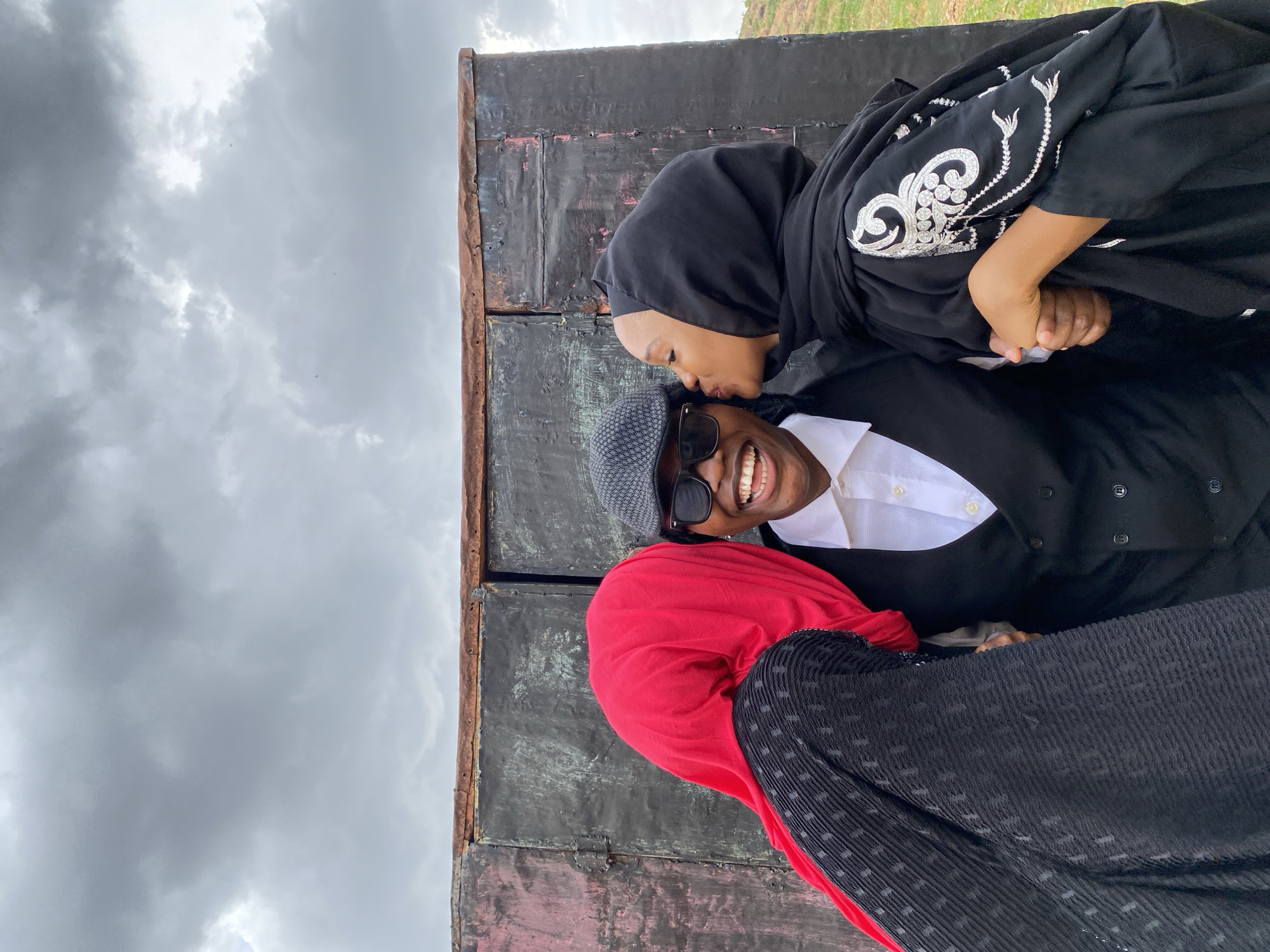 A photograph depicts three people in front of a metal structure against a cloudy sky and grassy land in the background.
One person, wearing a black headscarf and a black garment with white embroidery, is kissing another person on the cheek. The second person is wearing a gray hat, sunglasses, a white collared shirt, and a black vest, and is smiling. The third person is wearing a red headscarf and a black pleated garment. The individuals appear to be embracing near what seems to be a rusted metal wall.
The photograph's setting is possibly in Gwong, Nigeria.