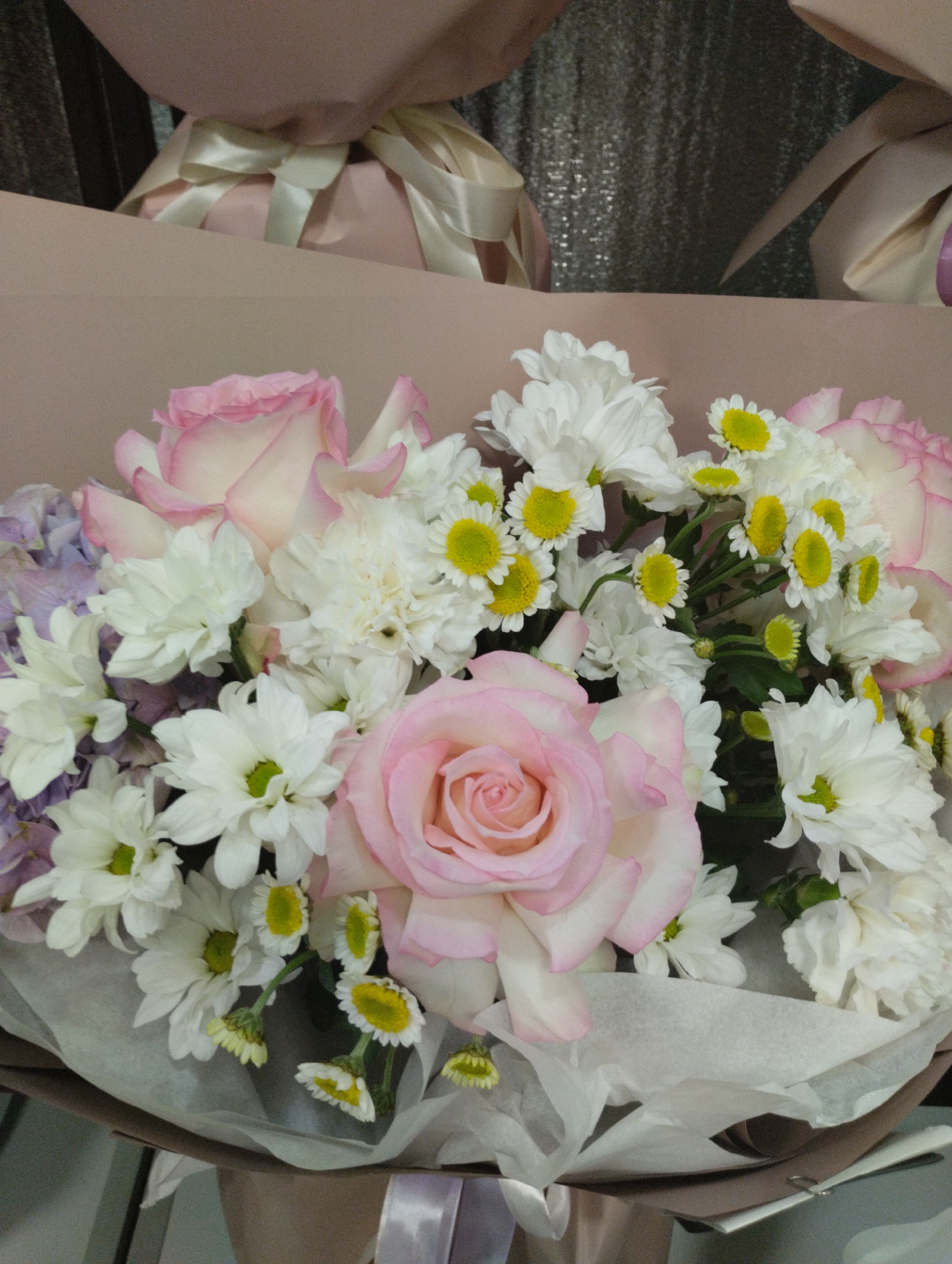 The image shows a bouquet of flowers, primarily pink roses and white daisies, arranged in a light brown paper wrapping. The bouquet is displayed in what appears to be a retail setting, with other wrapped gifts visible in the background. The overall aesthetic suggests a floral shop or a gift-wrapping service. There are no people or discernible actions in the image. The location in Kostanay, Kazakhstan, cannot be determined from the visual information provided.