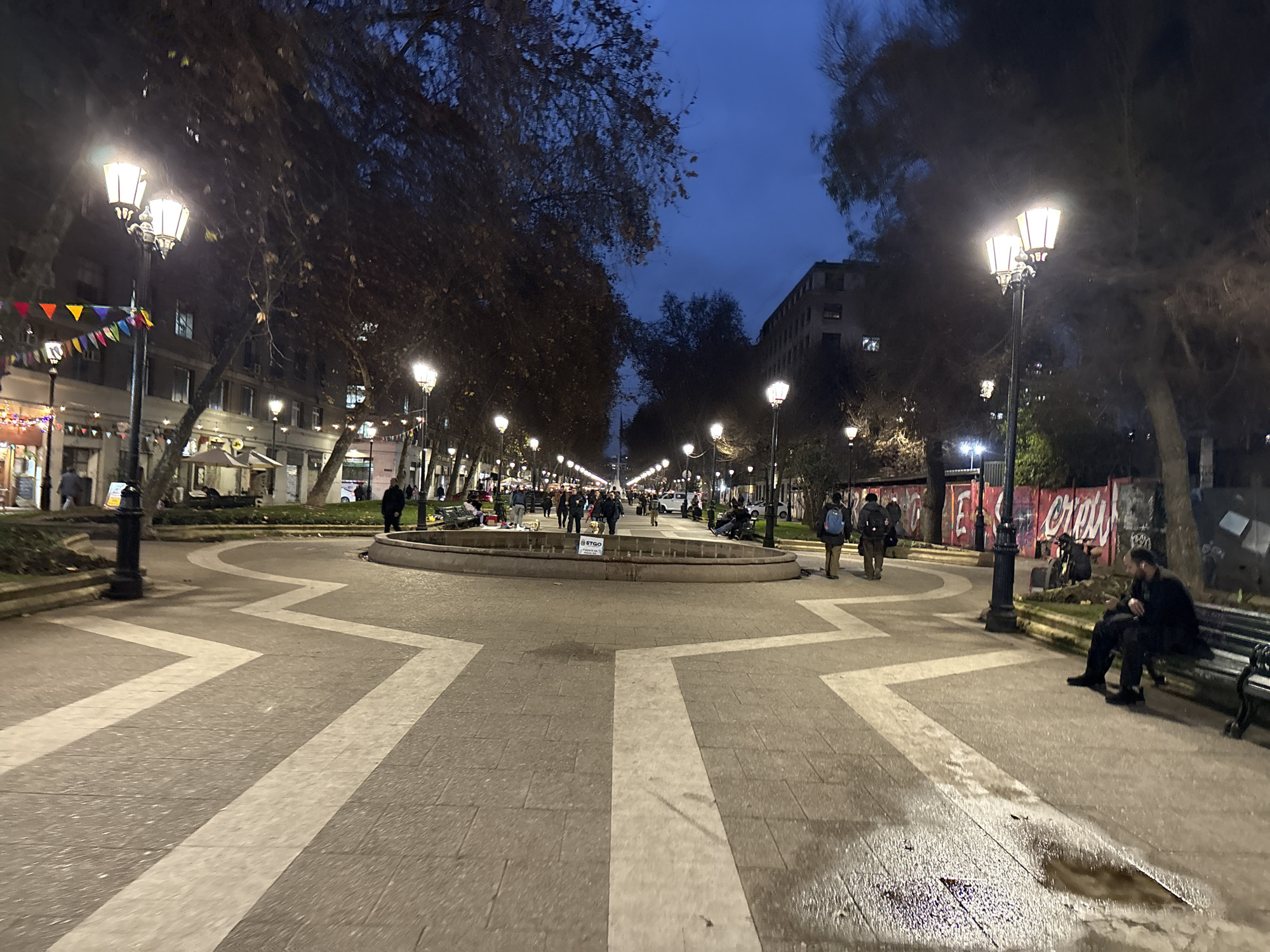 The image captures a view of Plaza Italia in Santiago, Chile at dusk. A wide, pedestrianized area with a fountain at its center is illuminated by streetlights and building lights. The ground is paved with stone and features a distinctive white zigzag pattern. Several trees line the sides of the plaza, their bare branches reaching towards the darkening blue sky. A number of people are seen walking, sitting on benches, and standing around the fountain. A man in a black jacket and pants is seated on a bench on the right side of the frame, looking at his phone. On the left side, a man in a dark jacket and backpack is walking away from the camera. The buildings in the background are typical of an urban environment, with visible windows and illuminated interiors. The overall atmosphere is one of evening activity in a public urban space.