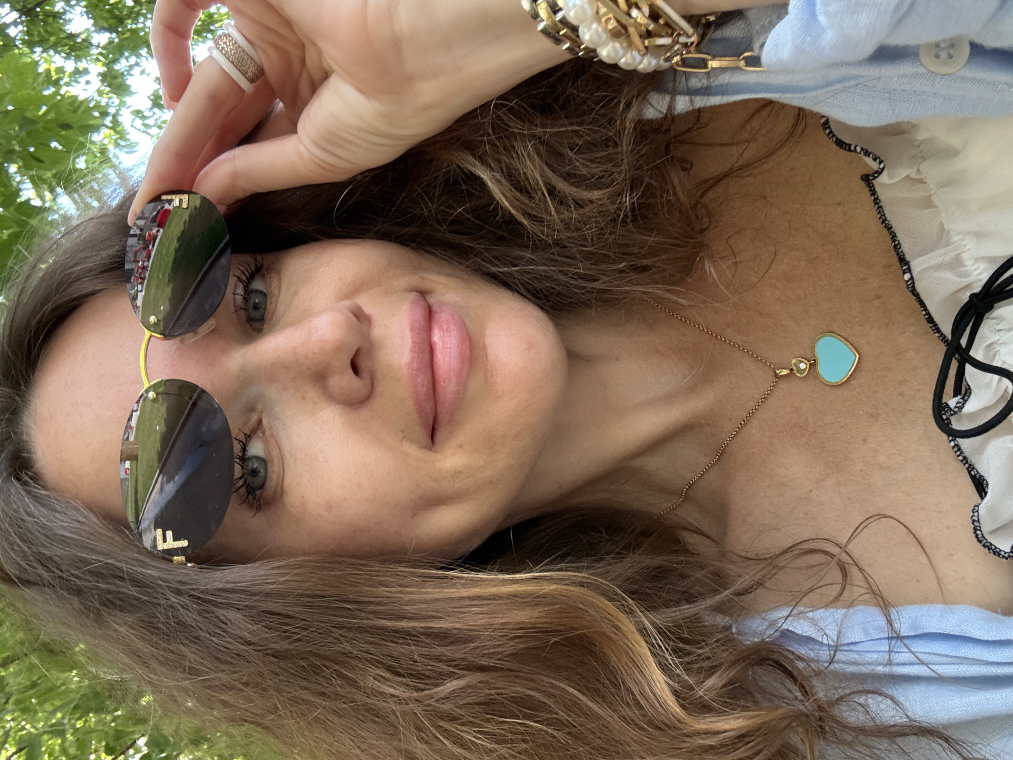 In a close-up, outdoor portrait, a fair-skinned woman with long, wavy brown hair adjusted her dark, oval sunglasses with a gold frame and an "F" logo. The sunglasses reflected a street scene with red vehicles and green grass. Her left eye, with dark lashes, was visible, and a dimple was present on her cheek. She wore a delicate gold necklace with a heart-shaped pendant containing a turquoise stone and a small diamond. On her right hand, she wore two rings, one with a textured rose gold band and the other a white band with a rose gold pattern. A stack of bracelets, featuring pearls and gold chains, adorned her right wrist. She was dressed in a light blue collared shirt over a white garment with black trim. The background consisted of blurred green foliage, suggesting an outdoor setting in a park or garden, possibly in Moscow, Russia, given the presence of city infrastructure reflected in her sunglasses.