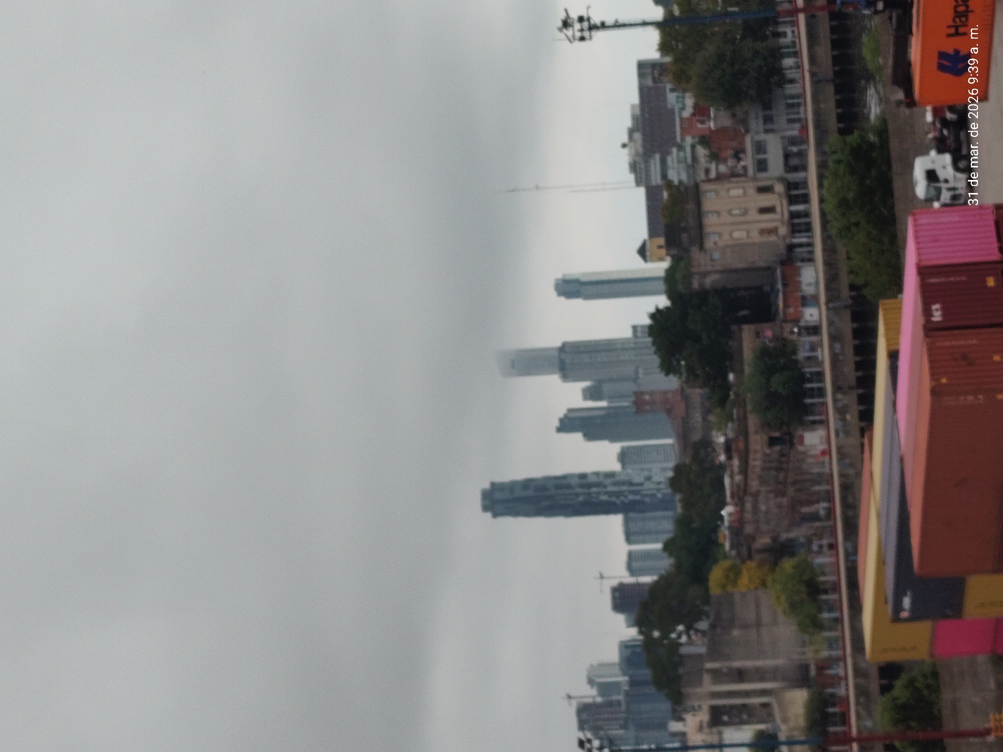 The image captures an overcast day in Dock Sud, Argentina. In the foreground, stacks of colorful shipping containers are visible, suggesting a port or industrial area. Behind the containers, there's a waterway lined with buildings and vegetation. In the distance, the skyline of a city, possibly Buenos Aires, is partially obscured by clouds and fog. The urban landscape includes a mix of low-rise and high-rise buildings. The date "31 de mar. de 2026 9:39 a. m." is imprinted on the image. There are no people visible in the shot. The primary focus is on the juxtaposition of industrial infrastructure and the urban environment against a backdrop of heavy weather.