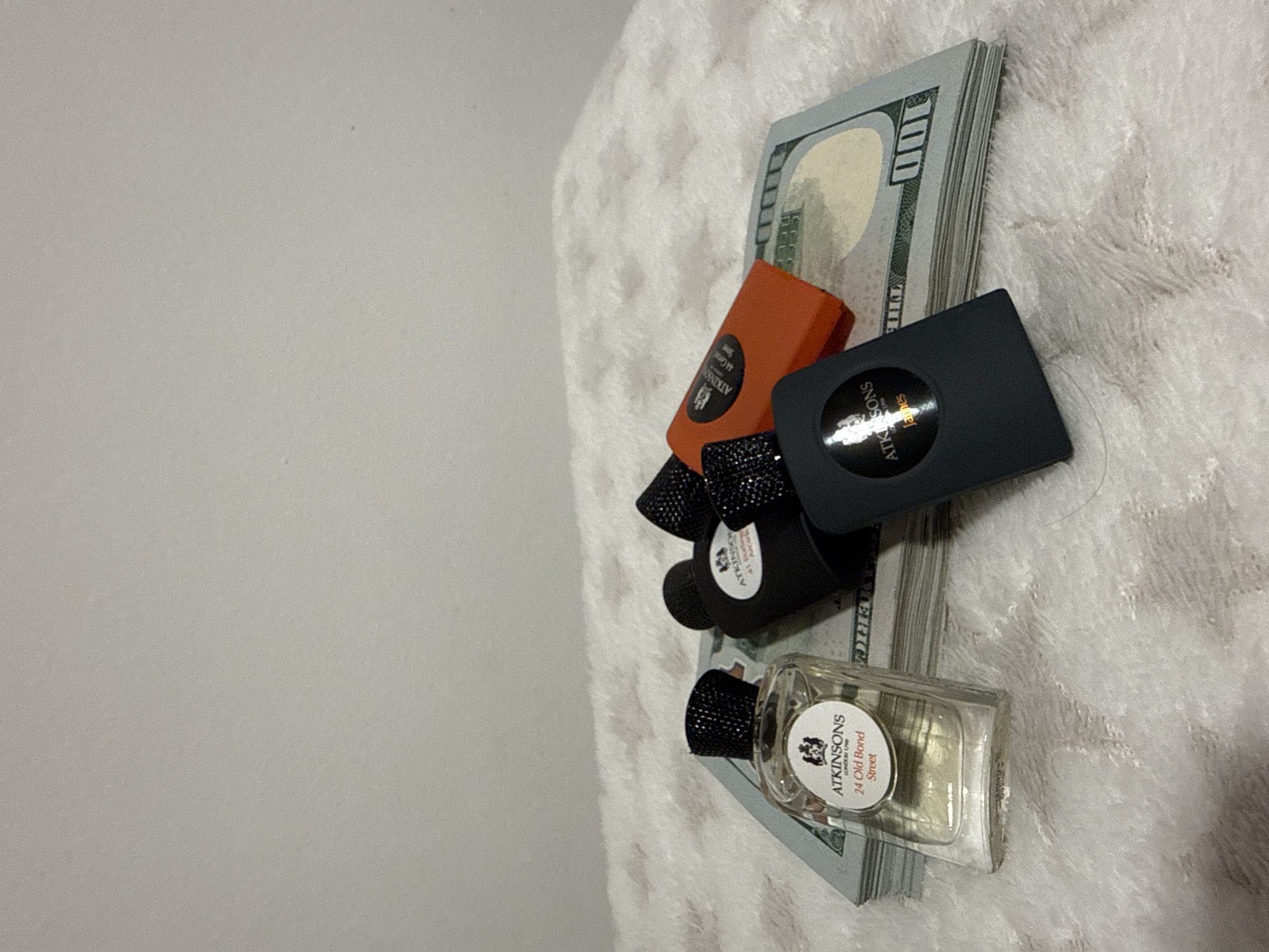 The image shows a stack of US one hundred dollar bills lying on a textured white surface. On top of the money are four perfume bottles or vials from the brand ATKINSONS. The bottles are of different shapes and sizes and include a rectangular bottle labeled "24 Old Bond Street". The background is a light gray wall. The location cannot be determined from this image.
