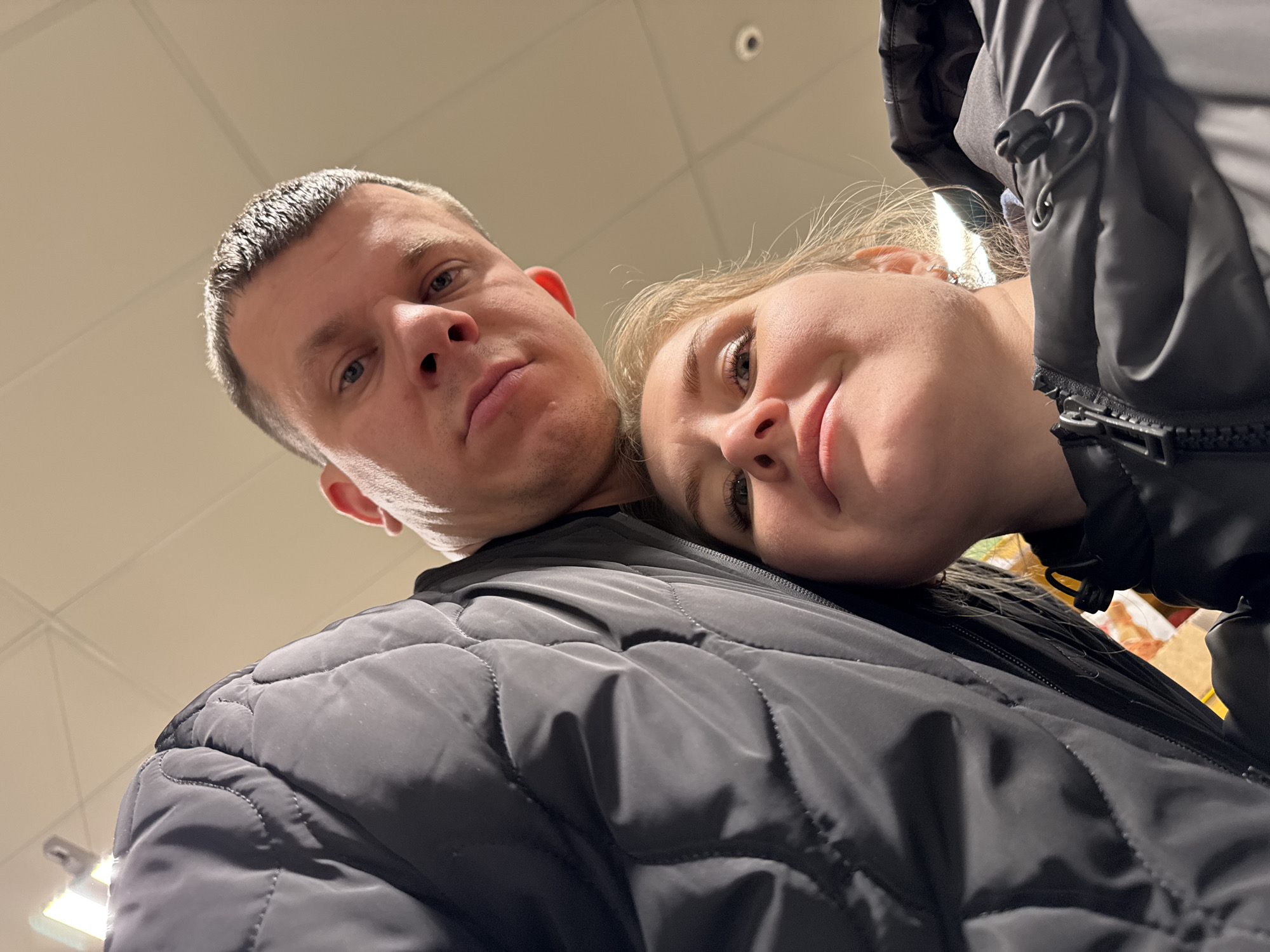 A low-angle selfie captures a man and a woman together in what appears to be a retail environment. The man, with short, light-colored hair, is to the left, his expression neutral. To his right, the woman rests her head on his chest. Both are wearing dark, quilted jackets. The background suggests the interior of a store, with ceiling lights and merchandise visible. It is difficult to confirm the location of Горный, Russia without further context or visual clues.