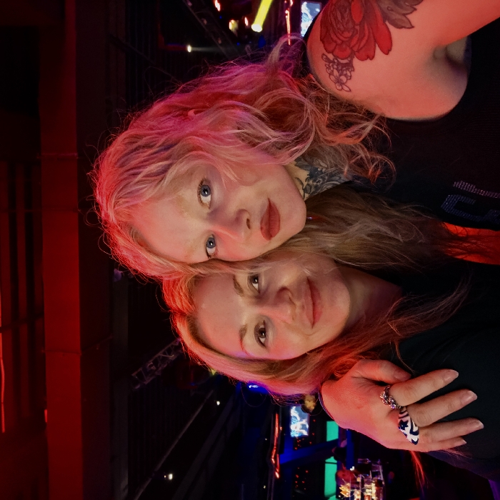 The scene captures two adult females posing for a photograph inside a dimly lit entertainment venue in Saint Petersburg, Russia. The individual on the left possesses blonde hair, is smiling faintly, and wears a dark garment. Her right arm is positioned around the shoulders of the person on the right. A prominent white ring with dark blue abstract patterns and a silver-toned band is visible on her right ring finger. The individual on the right features blonde, wavy hair, illuminated with a red hue, fair skin, light-colored eyes, and light-colored eyebrows. She has red lipstick applied and displays extensive tattooing, including dark floral motifs on her left shoulder and intricate patterns on her neck. She wears a dark, embellished tank top. Both subjects maintain direct eye contact with the camera. The background reveals structural trusses, multiple colored lights (predominantly red, with distant blue, green, and yellow accents), and indistinct lounge-style seating, consistent with a modern nightlife establishment.