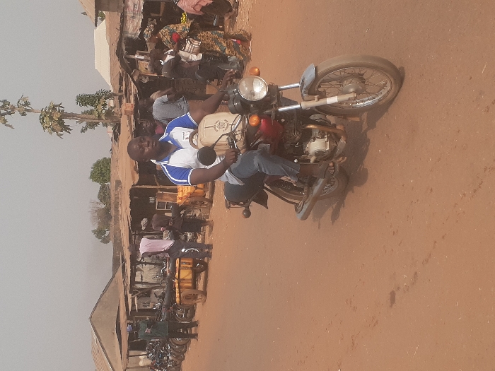 An individual is operating a red and black motorcycle on an unpaved, reddish-brown dirt road in Gwadanye, Nigeria. The driver, a Black male wearing a white and blue polo shirt, blue jeans, and dark sandals, has a light-colored backpack on his right shoulder. He is looking slightly right, towards the background. The motorcycle features a circular headlight and spoke wheels.
To the left of the road, a line of several parked motorcycles is visible alongside rudimentary structures with corrugated metal roofs. Two males stand near these parked vehicles, one in a green shirt and blue cap, another in a light-colored shirt. A wooden pushcart, laden with yellow containers or goods, is positioned nearby.
To the right of the road, a bustling market scene unfolds with numerous individuals and makeshift stalls constructed with wooden frames and simple roofs. Market-goers, including women in brightly patterned clothing, are engaged in various activities. Another wooden pushcart with yellow contents is present further back. Tall, slender trees with sparse foliage punctuate the background behind the market, set against a hazy, light grey sky.
