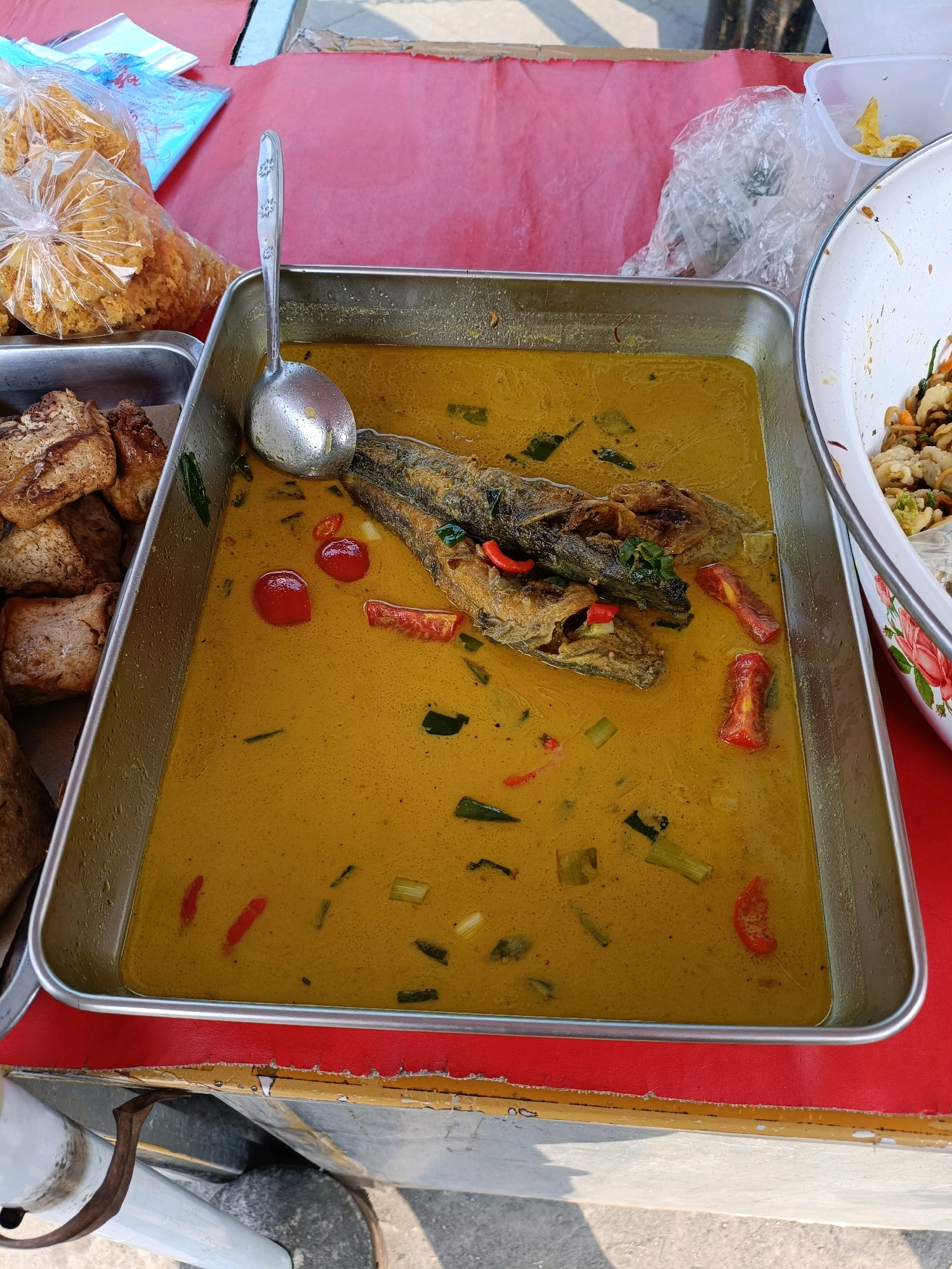An overhead shot shows a large metal tray filled with a yellow curry containing a whole fried fish, red chili peppers, and green vegetables. The tray sits on a red surface, possibly a table or a stall. To the left of the tray, a smaller metal container holds several pieces of fried tofu. In the background, plastic bags of what appear to be fried snacks are visible. To the right, a portion of a white bowl with a floral pattern is visible, containing another dish. The overall setting suggests a food stall or market in Maguwoharjo, Indonesia.