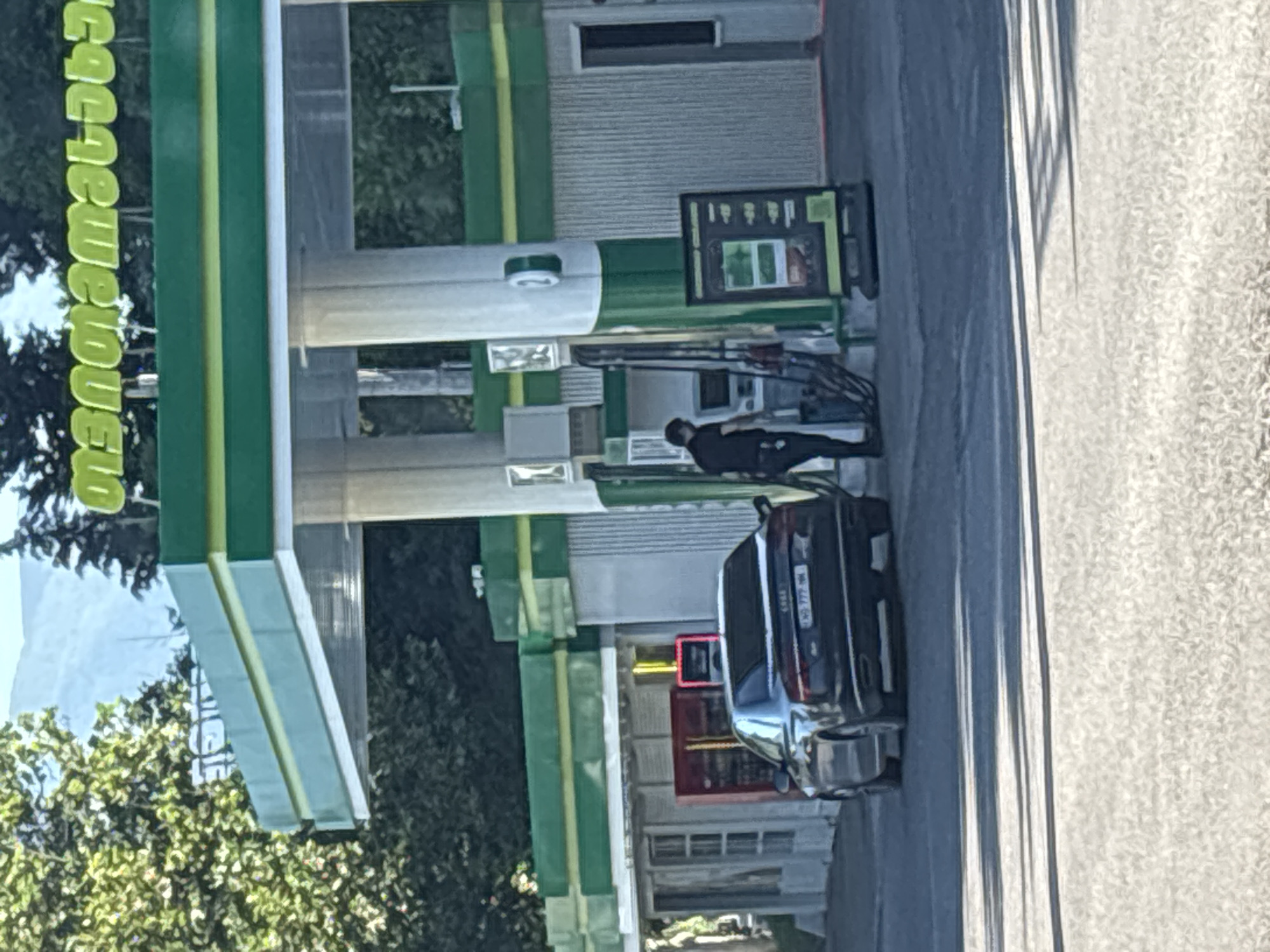 An Audi sedan, black, is parked at a gas station in Tbilisi, Georgia. The license plate is KG 777 NN. A man, wearing a black shirt and black pants, is standing at a gas pump, appearing to refuel the vehicle. The gas station has a distinctive green and white design. The background shows trees and mountains. A red vending machine is visible on the left side of the image.