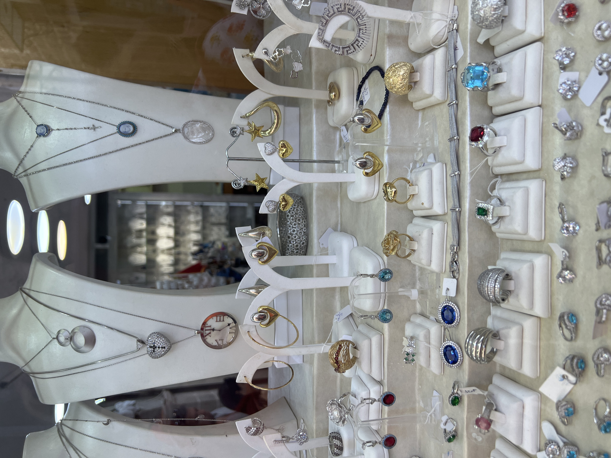 A window display of jewelry features necklaces, earrings, and rings. The jewelry is displayed on white mannequins and stands. The necklaces include a silver chain with a pendant of two interlocking circles, a silver chain with a heart-shaped pendant encrusted with clear stones and a black cross, and a silver chain with a round pendant resembling a clock face with Roman numerals. Other necklaces have pendants with blue and white cameo images, and a cross. Earrings on display include gold hoops, gold heart-shaped earrings, gold star earrings, silver teardrop earrings, and earrings with blue or red stones. Rings include a silver ring with red stones, a gold ring with red stones, a silver ring with a floral design, and a variety of silver rings with clear or blue stones. A beaded bracelet is also visible. The jewelry is arranged on a light-colored surface, and a patterned silver bracelet is displayed horizontally across the middle shelf. The background shows blurred images of other merchandise and lighting fixtures. The scene appears to be a jewelry store display.