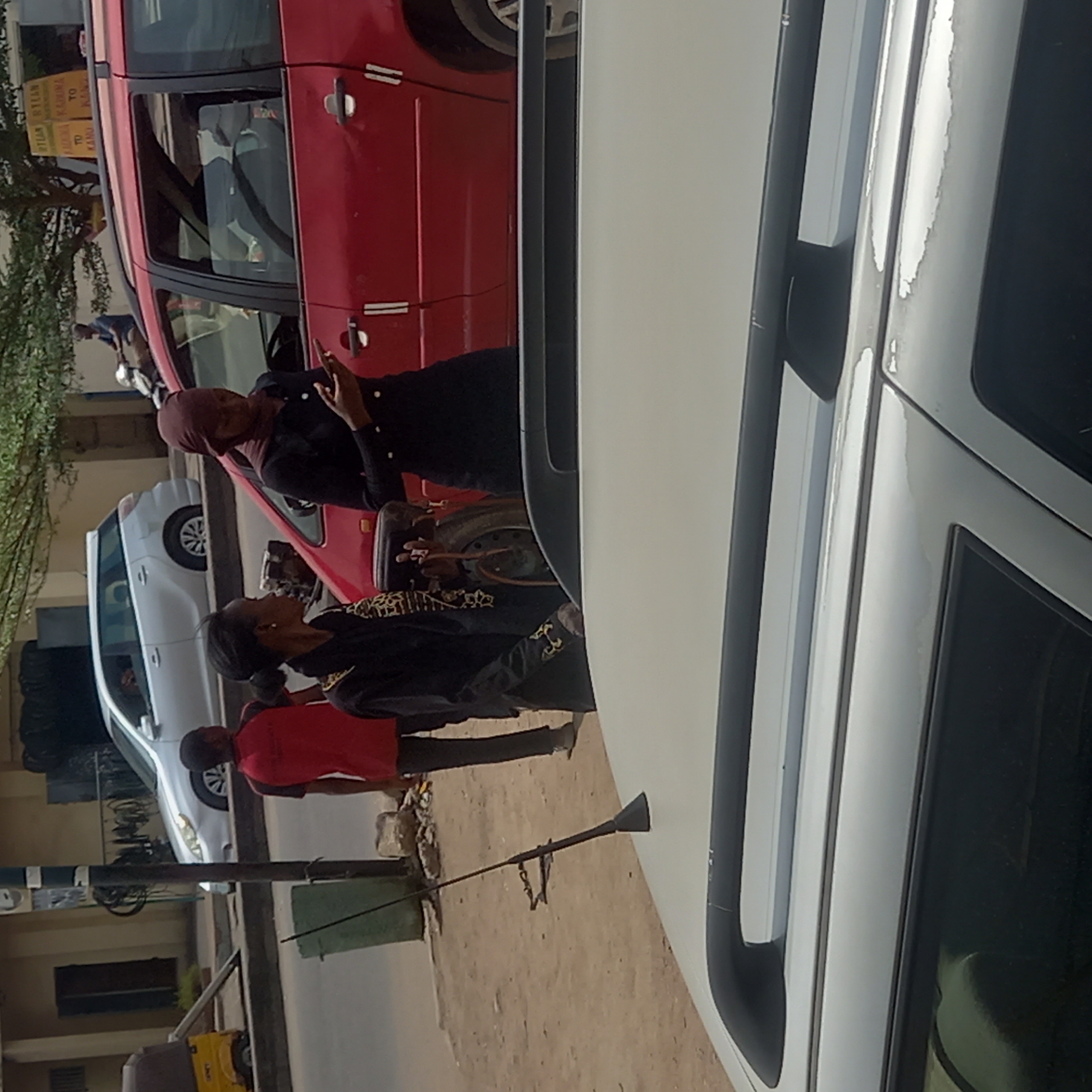 The image shows a street scene in Dadi Riba, Nigeria. In the foreground is the side of a white vehicle. A red van is parked adjacent. Two people are standing by the red van. One wears a maroon head covering and black clothes and is gesturing. The other person wears black clothes with gold embellishments. Behind them, a person in a red shirt stands near a gray car. There is a trash can and a slender pole with a tool hanging from it on the side of the road. A building is visible in the background.