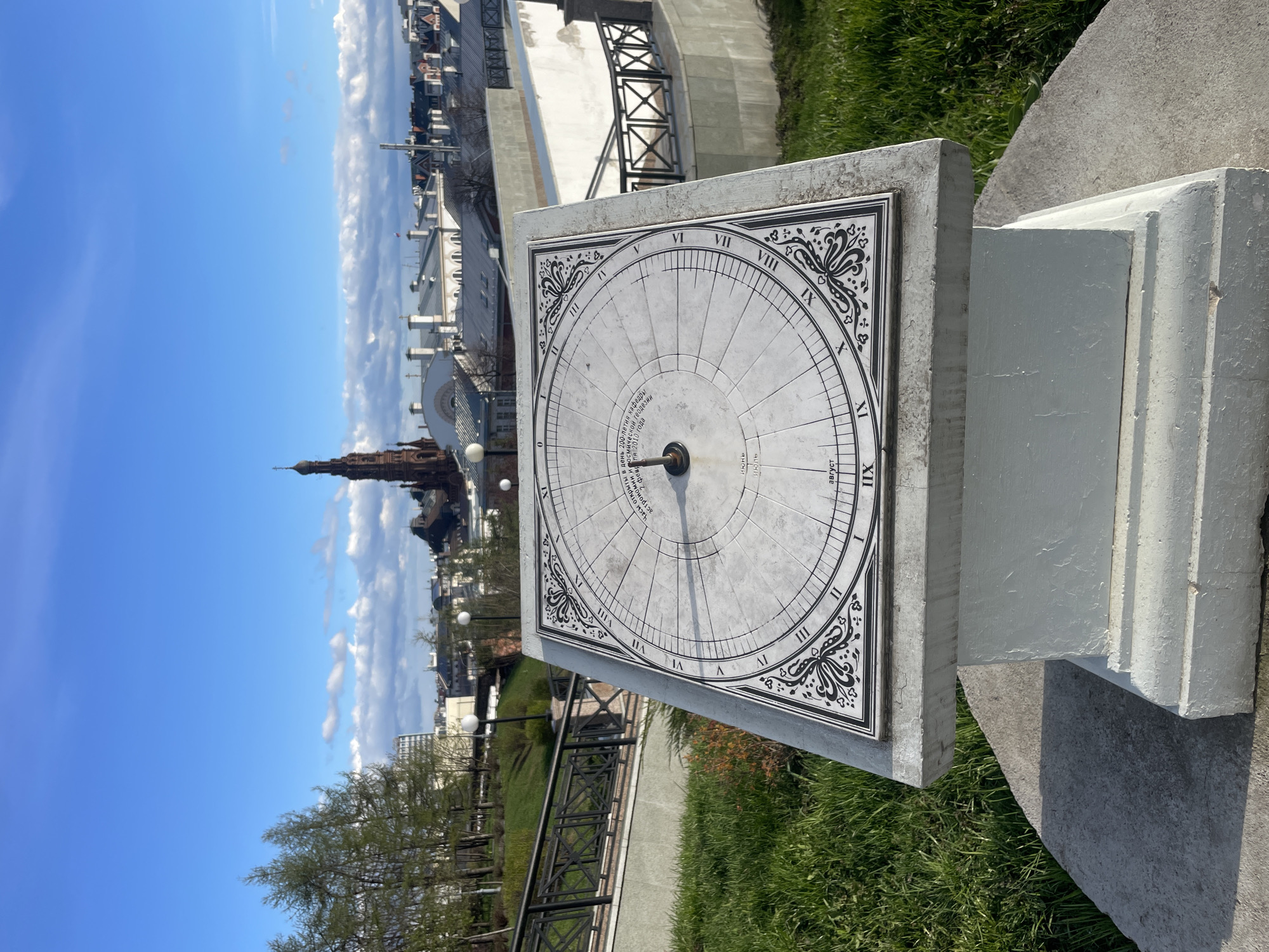 The image captures a horizontal sundial set in an outdoor, elevated location in Kazan, Russia, under a bright, partly cloudy blue sky. The central subject is a large, square sundial made of light-colored stone or concrete, with its surface marked by black Roman numerals (I through XII) indicating hours. Decorative flourishes adorn the corners of the sundial's face, and a central gnomon casts a shadow, although the precise time is not clearly discernible.
Significant Cyrillic text is inscribed on the sundial. According to the OCR, it reads: "Часы открыты в день 200-летия кафедры астрономии и космической геодезии 2 февраля 2011 года." This translates to: "These clocks were opened on the 200th anniversary of the Department of Astronomy and Space Geodesy on February 2, 2011." This detail anchors the sundial's location within an academic and historical context, likely related to Kazan Federal University. Month indicators like "Июль" (July) and "Август" (August) are also visible.
In the background, against the expansive blue sky, a distinctive red-brick tower with a spire, characteristic of Kazan's architecture, rises above other city buildings. The foreground and surrounding area feature vibrant green grass, hinting at spring or early summer, a pathway, and a black metal fence. The scene is illuminated by bright daylight, suggesting a sunny day. No people are present, lending a tranquil atmosphere to this blend of historical architecture and scientific monument.