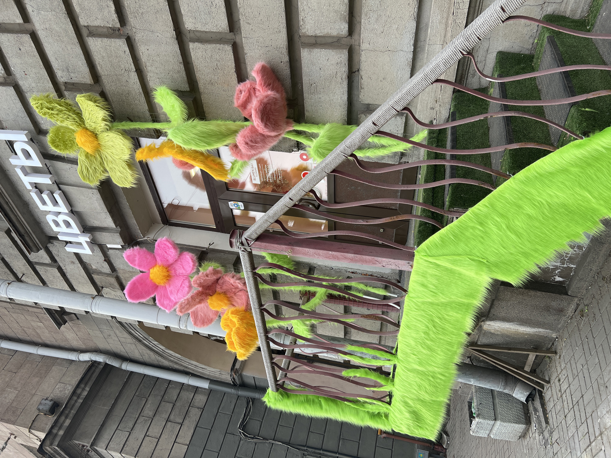 The image shows the exterior of a flower shop located in what appears to be Saint Petersburg, Russia. The shop's name, "Цветы" (Tsvety), meaning "Flowers," is displayed in white block letters above the entrance. Large, colorful, plush flowers in shades of yellow, pink, and orange decorate the exterior wall. The metal railing of the staircase is wrapped in bright green faux fur. The stairs are covered with artificial turf. The architecture of the building suggests a historic urban setting, with brick and stone structures visible. A drainpipe runs along the side of the building. The image appears to be taken from a low angle, looking upwards towards the shop.