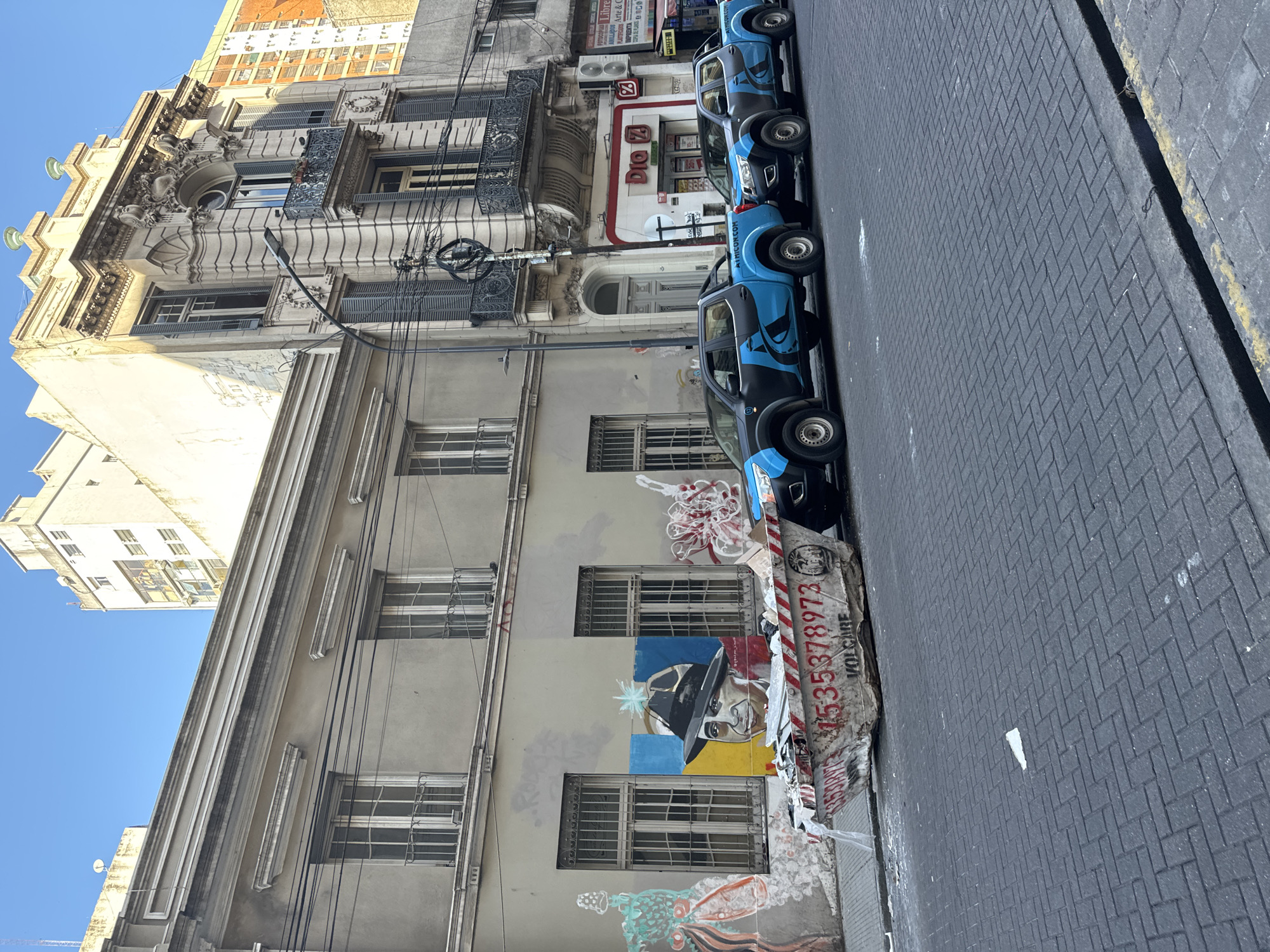 The image shows a street scene in Buenos Aires, Argentina. The view is taken from street level, looking along a row of parked blue and black pickup trucks. The trucks are parked along a building with graffiti on the wall. The building appears to be in disrepair and has multiple windows with security bars. Above this building is another building with ornate architectural details, including balconies and decorative molding. Adjacent to the building is a store front with a sign that reads "Dio Z". The street is paved with gray bricks. The sky is visible in the upper left corner, and is a clear blue.