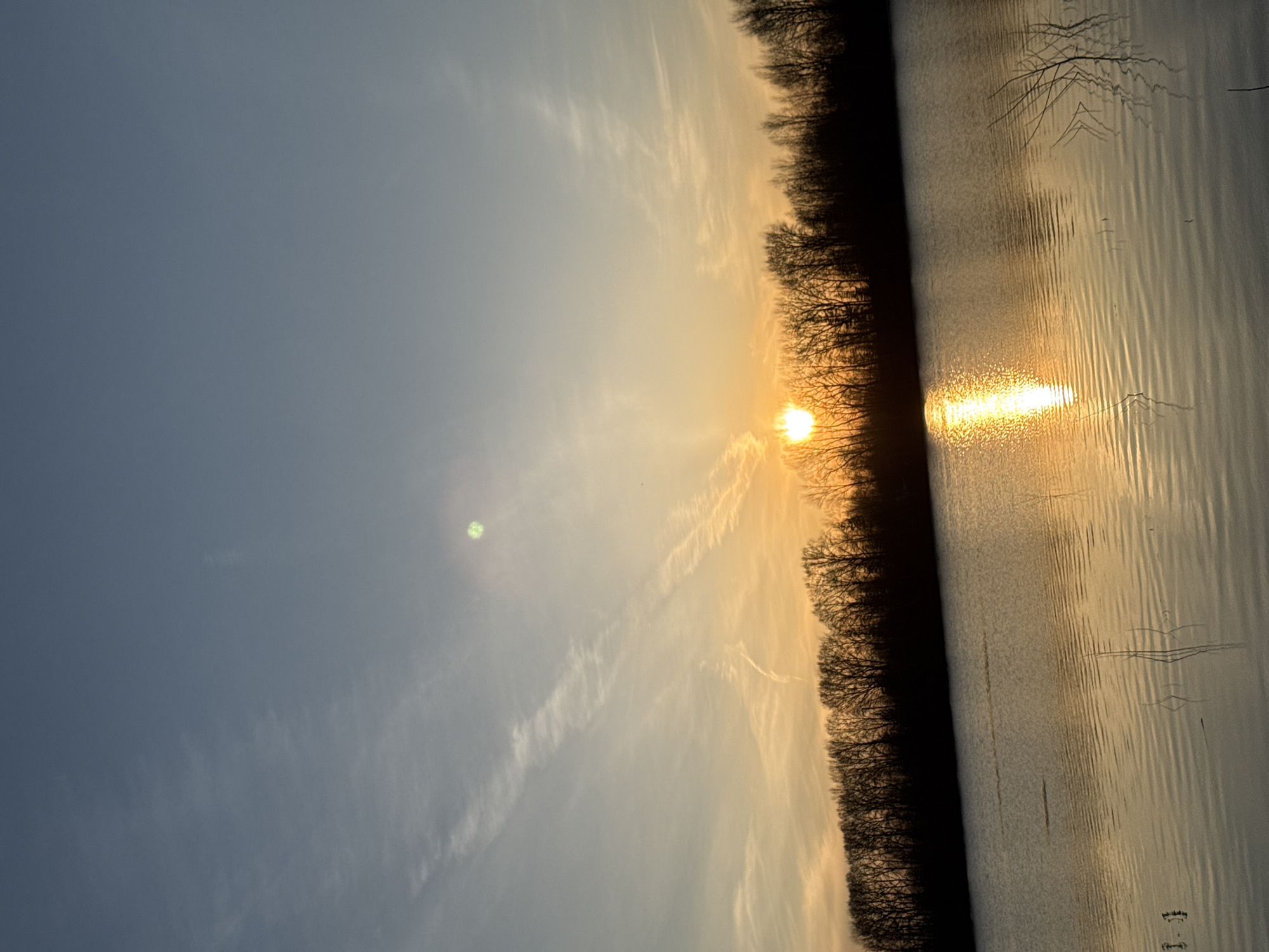 The image depicts a horizontal view of a sunset over a body of water, potentially a river or lake, in Chaikovsky, Russia. The sun is setting near the horizon, casting a bright reflection on the water. A line of trees is situated between the sun and the water, creating a silhouette effect. The sky has a gradient of colors, from light blue to orange, with visible contrails. The water's surface has ripples, indicating a slight breeze. There are no people visible in the image.