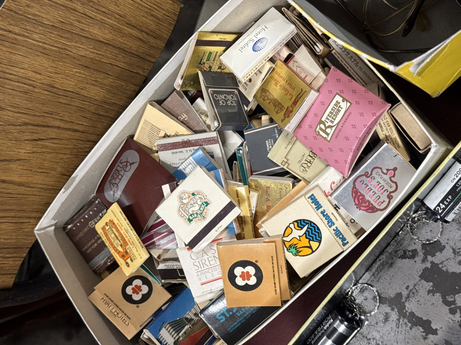 An overhead, slightly angled photograph displays an open, rectangular, off-white cardboard box filled with an unorganized collection of numerous matchbooks. The matchbooks, varying in color, size, and design, feature advertisements for various establishments such as hotels, resorts, casinos, and restaurants, with visible names including "Hyatt Hotels," "Americus," "Pacific Shore Hotel," "Pueblo Joe's," "Riverside Resort," "La Casita Marina," "Hotel Sofitel," and "Top of Toronto." Prominent logos and textual details are visible across many of the matchbook covers. Positioned outside the box, on the lower left, are two 35mm film canisters, each attached to a metallic keyring. One canister is labeled "KODAK PORTRA 400" and another indicates "24 EXP." The box and film canisters rest on a dark, textured surface, possibly a tabletop. A dark, non-descript object, potentially fabric, is partially visible along the far left edge. The background consists of a light brown, wood-grained surface, extending diagonally from the upper left. This scene is located in the city of Cagliari, Italy.