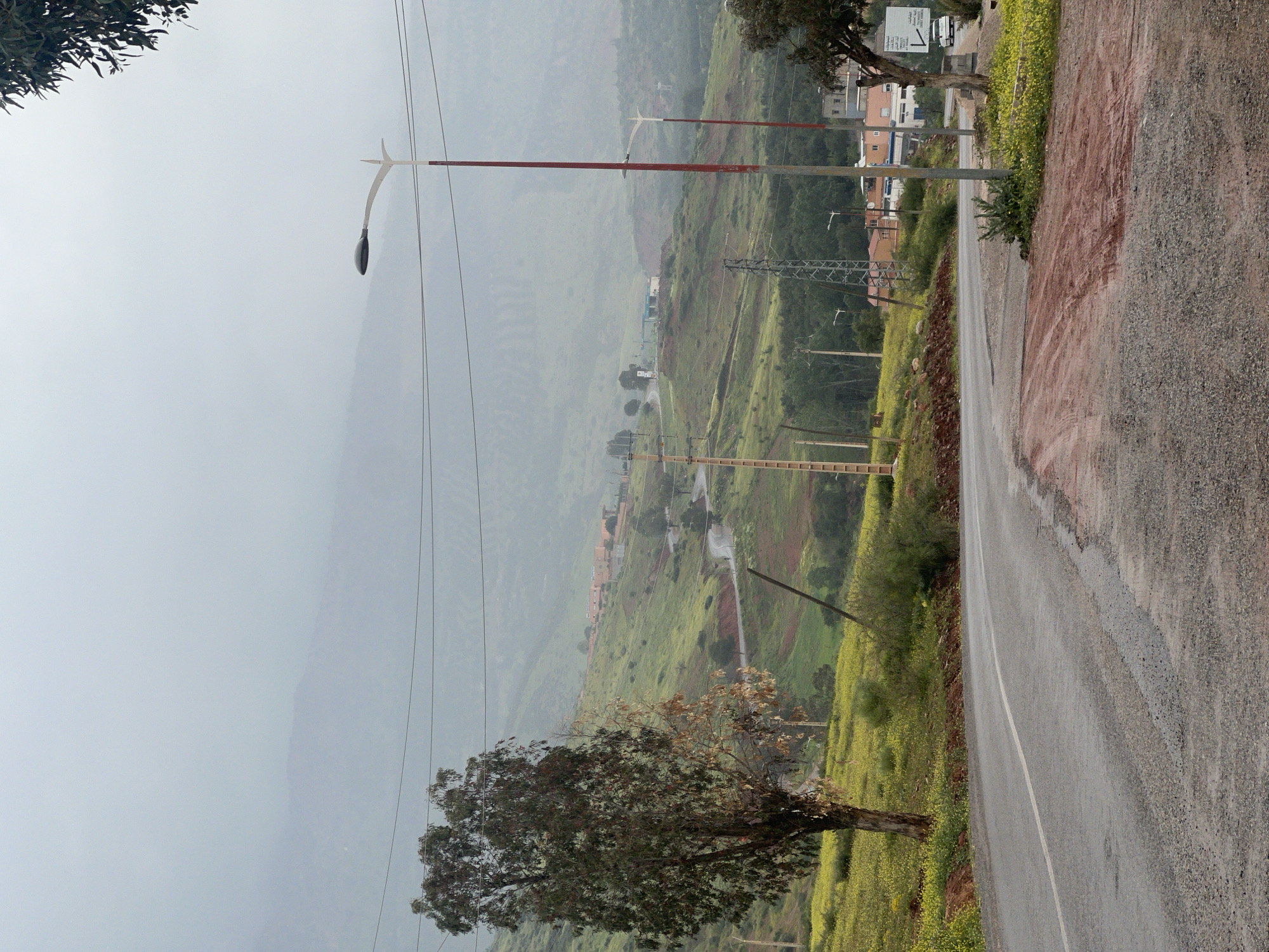The image shows a rural scene near Ouaouizert, Morocco. A paved road curves through a green, hilly landscape, dotted with yellow flowers and scattered trees. Utility poles and power lines are visible, intersecting the landscape. In the background, the terrain ascends into misty mountains. The road is wet, suggesting recent rain. Buildings are sparsely located in the distance along the hillsides.