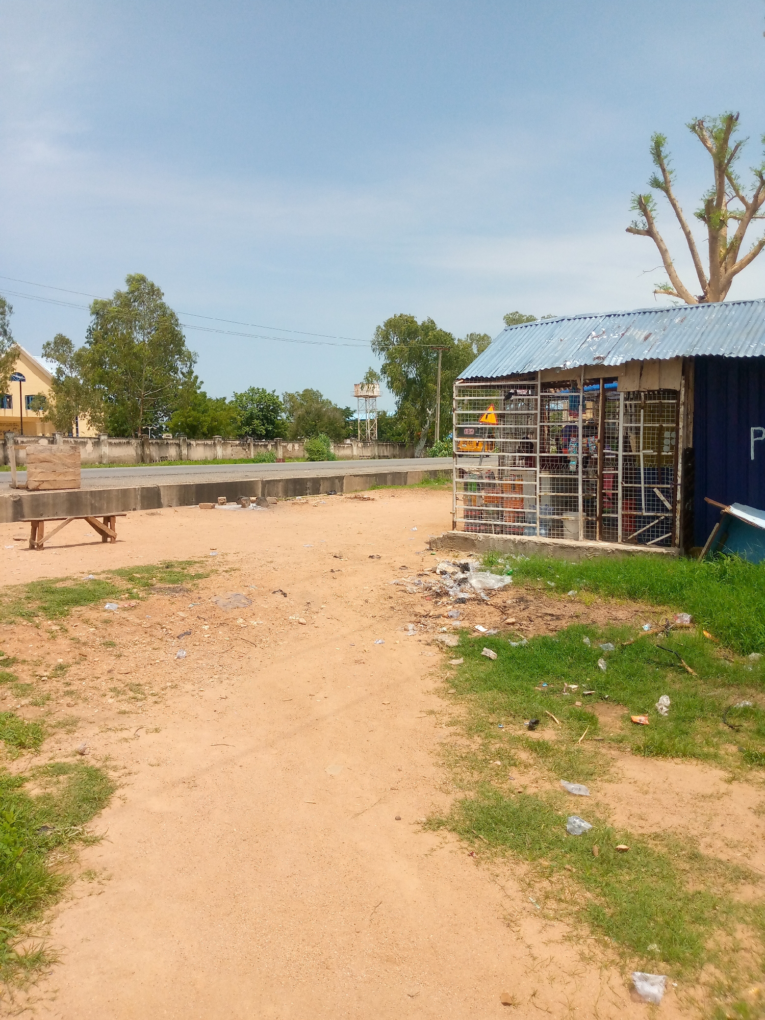 The image depicts a street scene in Jalingo, Nigeria. In the foreground, a sandy path is visible, with patches of green grass and scattered debris. A wooden bench sits to the left, adjacent to a stack of wooden planks. A small, open-air shop with a corrugated metal roof is situated on the right side of the frame. The shop is constructed with metal bars and contains various items for sale, including bottles and other goods. Behind the shop, a blue structure is partially visible with the letter "P" in white. A road runs horizontally across the middle ground, bordered by a concrete barrier. In the background, a wall separates the road from a building with a yellowish facade and blue windows. Trees are scattered throughout the scene, and a water tower is visible in the distance. The sky is clear and blue with some wispy clouds.