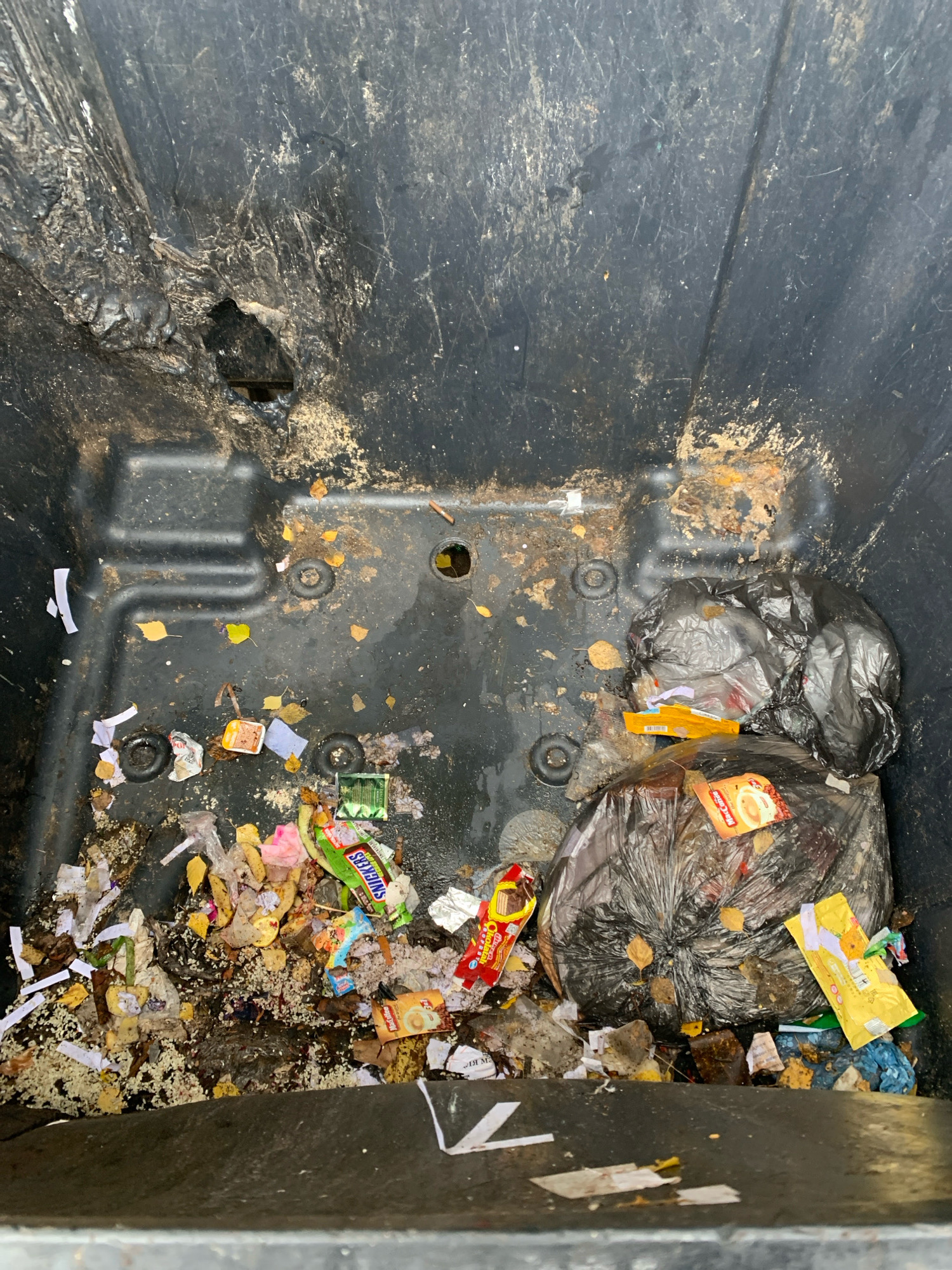 This image shows the inside of a black garbage bin, located in the city of Arkhangelsk, Russia. The bin is filled with various types of refuse, including crumpled paper, food wrappers, banana peels, and plastic bags. Among the debris, one can identify a "Snickers" candy bar wrapper, a yellow package with an image of coffee, and another yellow package with text and graphics that are not clearly legible. Two large black trash bags are visible, one partially obscuring the other. The bin itself is dirty and shows signs of wear, with scuff marks and a small hole in the upper left interior wall. Leaves and small pieces of debris are scattered throughout the bottom of the bin. The lighting suggests an outdoor setting.