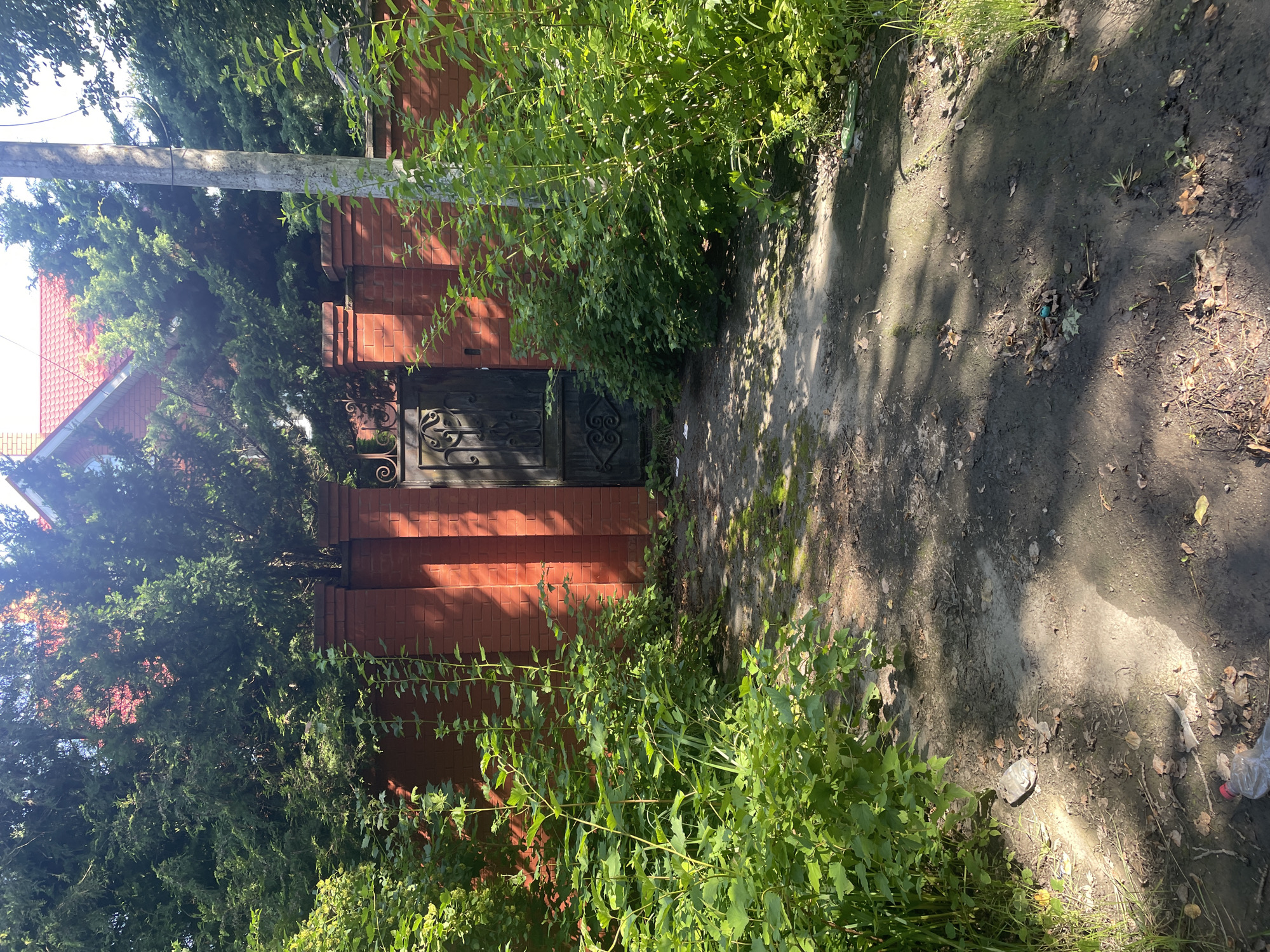 A dirt path leads to a black metal gate set within a red brick wall. The gate is ornate with decorative scrollwork. Lush green foliage borders the path and wall, with trees and a portion of a red-roofed building visible behind the wall and trees. A concrete utility pole stands to the right of the gate. The scene is illuminated by sunlight, casting shadows on the path. Debris, including plastic bottles and leaves, is scattered on the ground along the path and at the base of the wall.