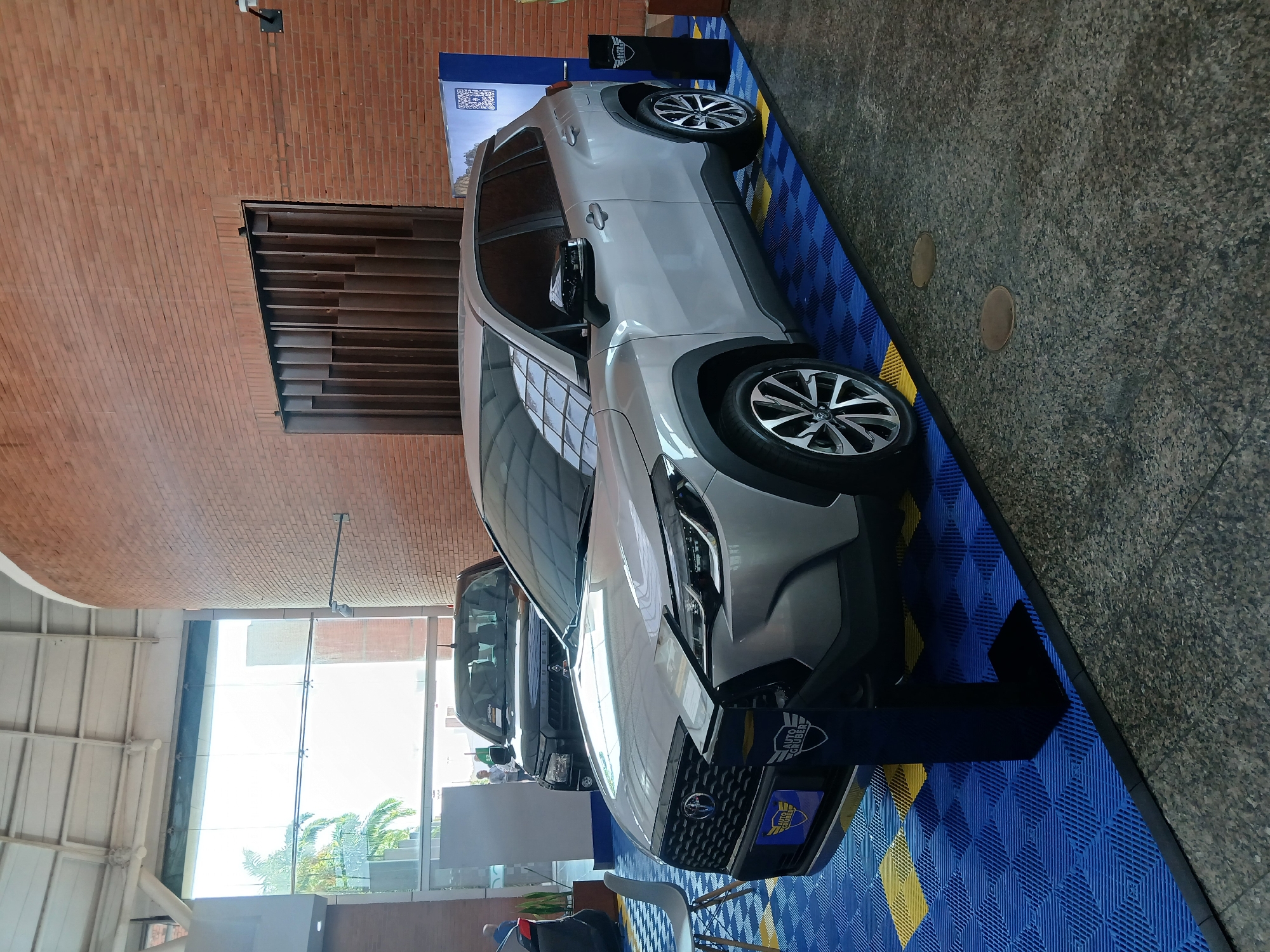 A silver SUV, possibly a Toyota Corolla Cross, is displayed on a blue and yellow tiled platform indoors. Another dark SUV is visible behind it. The location appears to be a car dealership in Ciudad Guayana, Venezuela, as evidenced by the "AUTO GRUBER" emblem on the displayed vehicle. Brick walls and large windows are visible, suggesting a showroom setting. No people are clearly visible, though there may be individuals in the background.