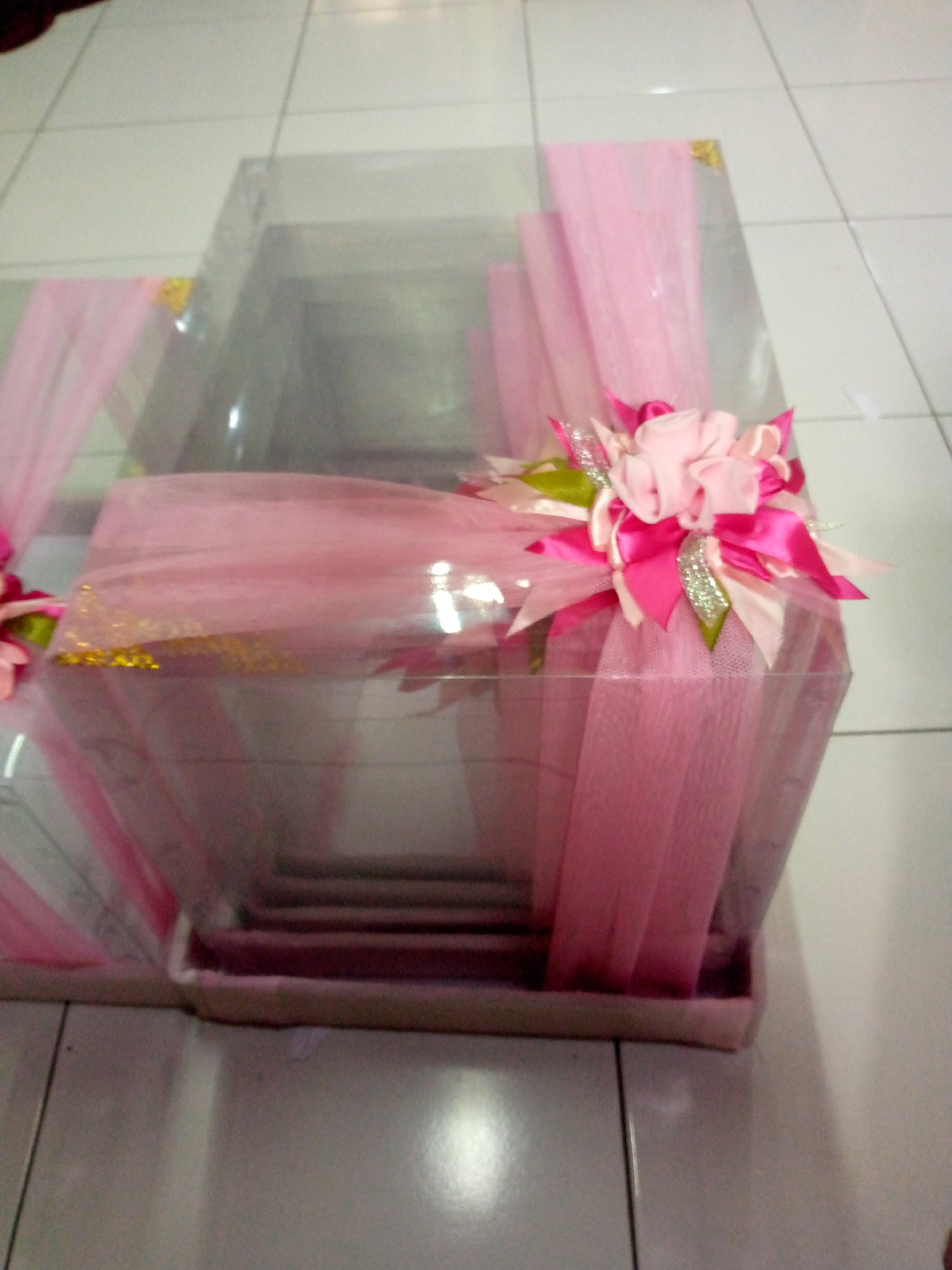 This is a close-up, high-angle, interior shot of a transparent gift box decorated with pink tulle and bows. The box is filled with layered items, possibly gifts, that are visible through the clear plastic. A decorative bow made of pink ribbons and glittery accents sits on top of the tulle, partially obscuring the contents. The gift box is placed on a tiled floor, with white and gray tiles visible. The lighting is bright, causing reflections on the plastic box and the floor. The scene appears to be set in a home or a shop in Sumberwono, Indonesia.