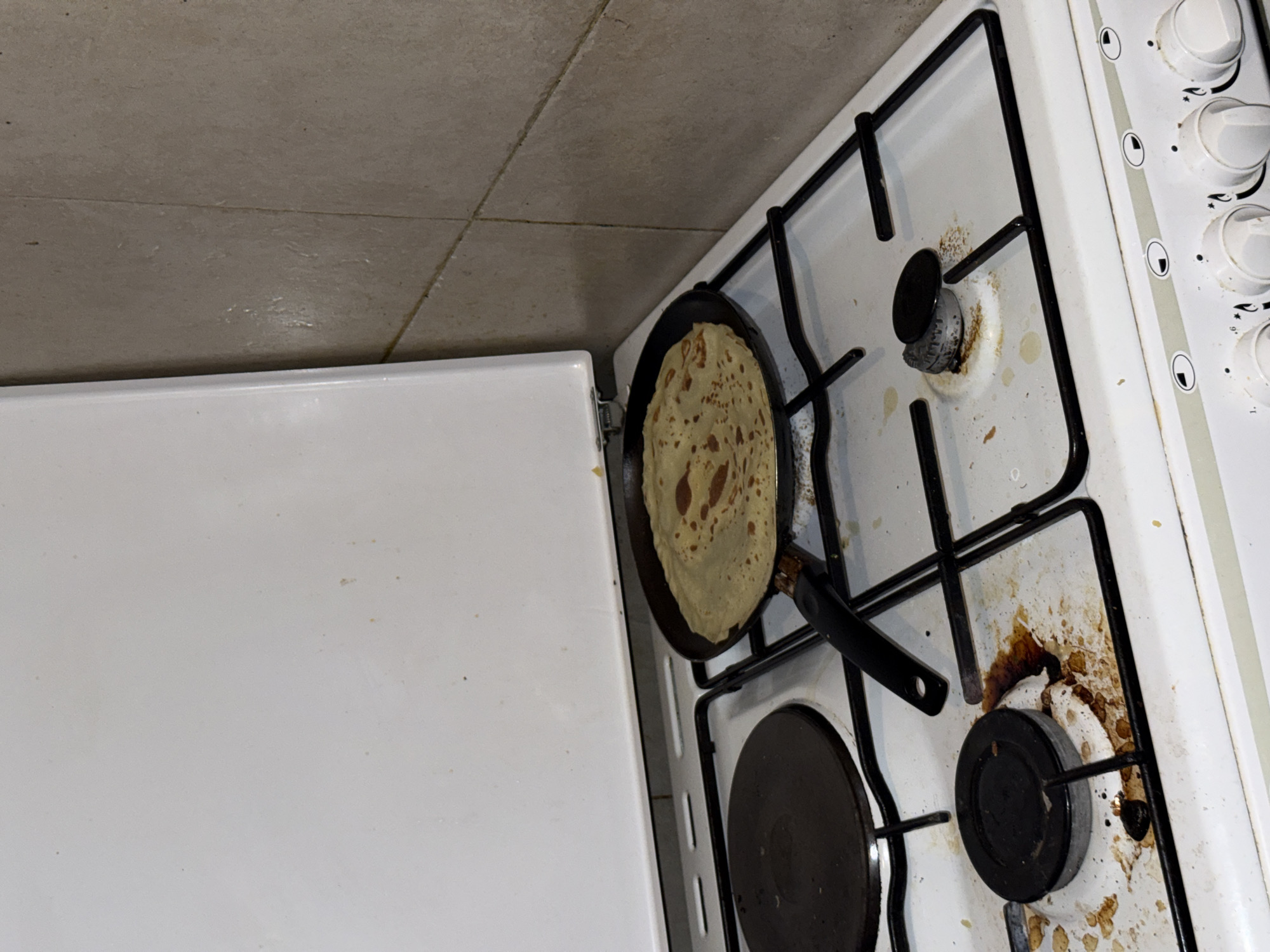 The image shows a top-down view of a white gas stove, likely located in a kitchen in Domont, France (based on image context). A black frying pan with a pancake inside rests on one of the burners. Another burner is visible below the pan. The stovetop shows evidence of cooking splatters and is in need of cleaning. A white surface, possibly a countertop or part of the stove itself, is visible on the left side of the frame. No people are visible in the image.