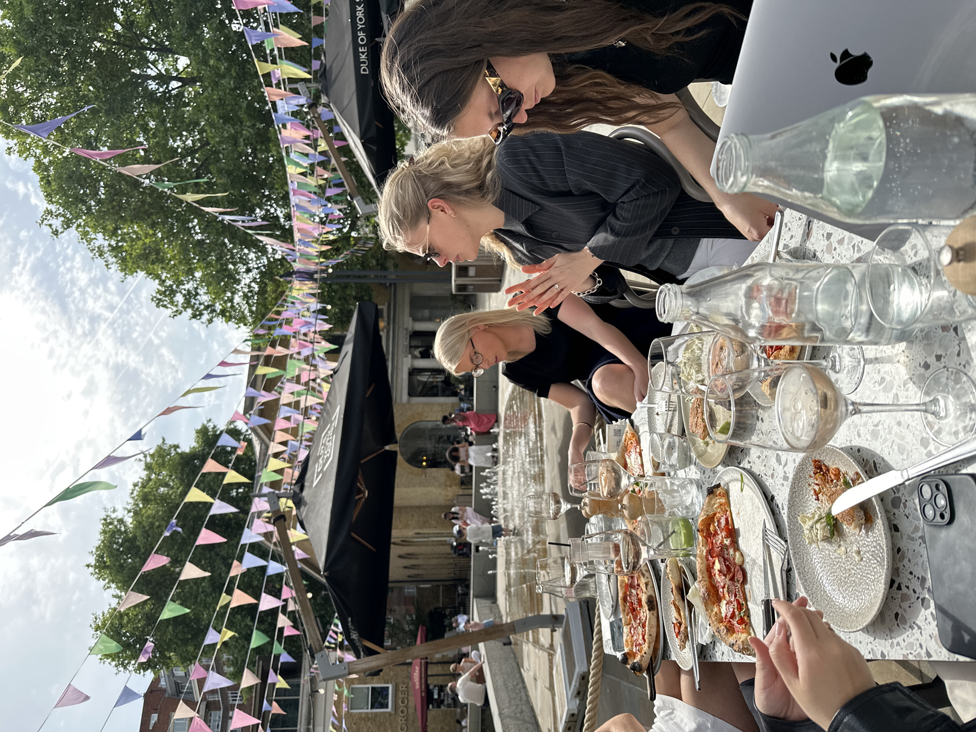 Three women are seated at a long table in an outdoor setting. The table is covered with plates of pizza, silverware, wine glasses, and water bottles. A laptop is visible on the table near the woman at the top of the frame. The women appear to be engaged in conversation. Colorful flags are strung overhead, and the surrounding environment includes trees and buildings. The sign "Duke of York Square" is visible, indicating the location as Duke of York Square.