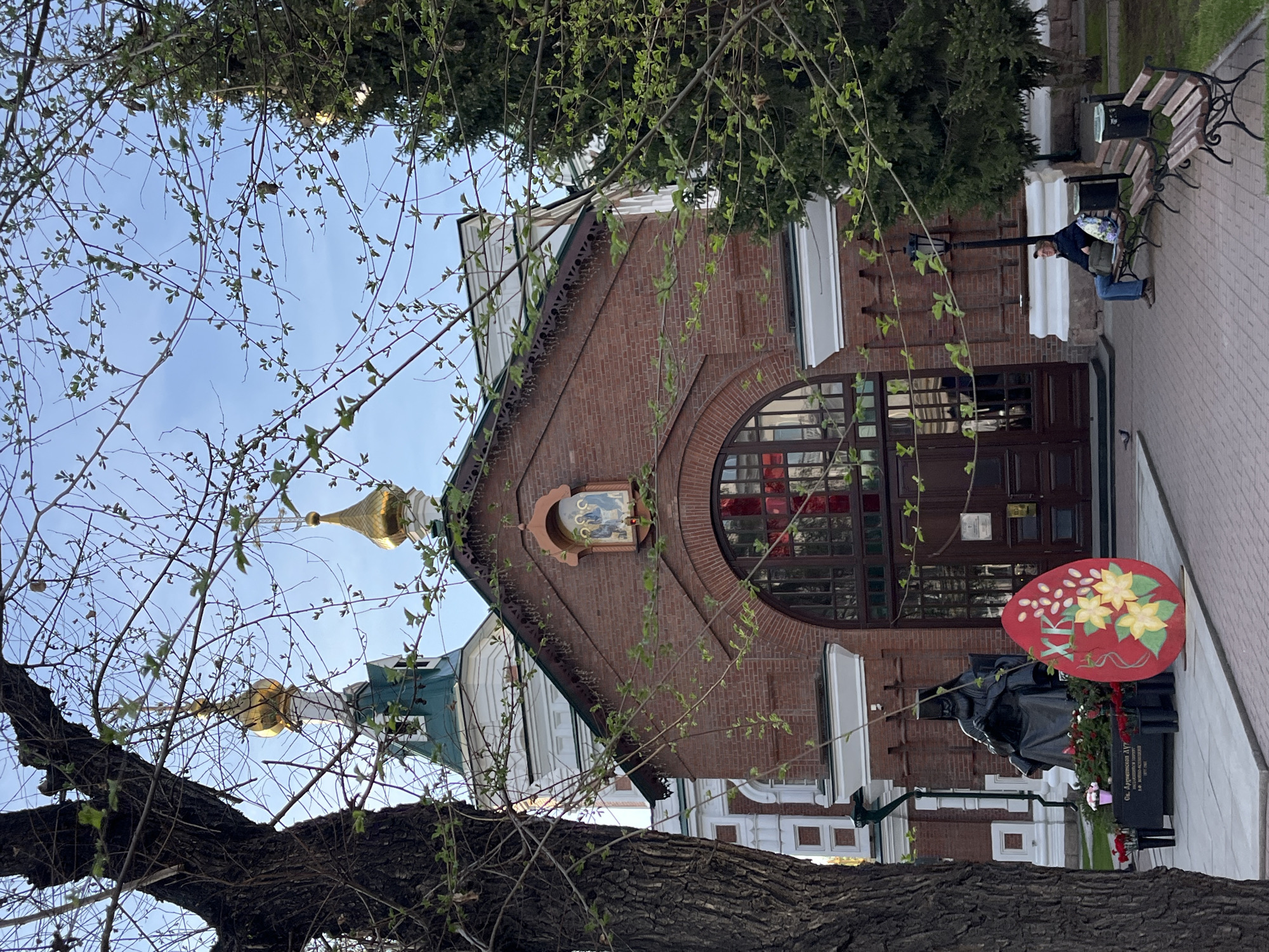 The image depicts a brick church building with a large arched window, topped with a small religious icon. The church also has a dome with a golden spire visible above and to the left. A large red Easter egg decoration sits in front of a statue near the entrance of the church. A bench and a person are visible in the background, on the right side of the frame. The scene appears to be in a city, potentially Unknown, Unknown. The image is framed by tree branches, obscuring some of the view.