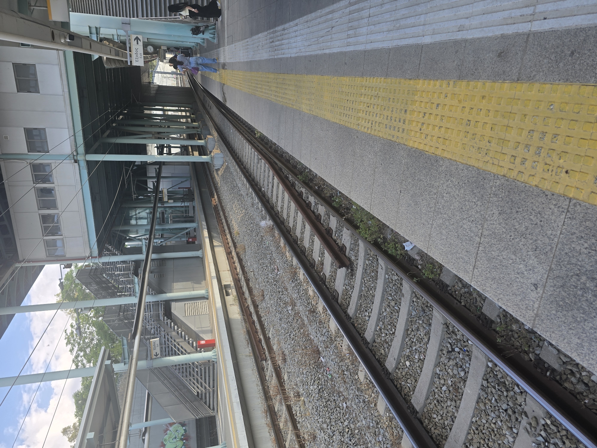 This is an eye-level, outdoor, medium-shot photograph taken at a train station. The scene depicts train tracks running diagonally from the lower left to the upper right of the frame. The tracks are surrounded by gravel and concrete. To the right of the tracks is a train platform with a yellow tactile paving strip along the edge. Beyond the platform, a building structure with blue supports and a white facade is visible. There are also stairs and an "Exit" sign in Greek and English. Two women are standing on the platform, and another person is visible further down the platform. The sky is blue with some clouds, and trees are visible in the background. The location is identified as the Agii Anargiri train station in Agii Anargiri, Greece.