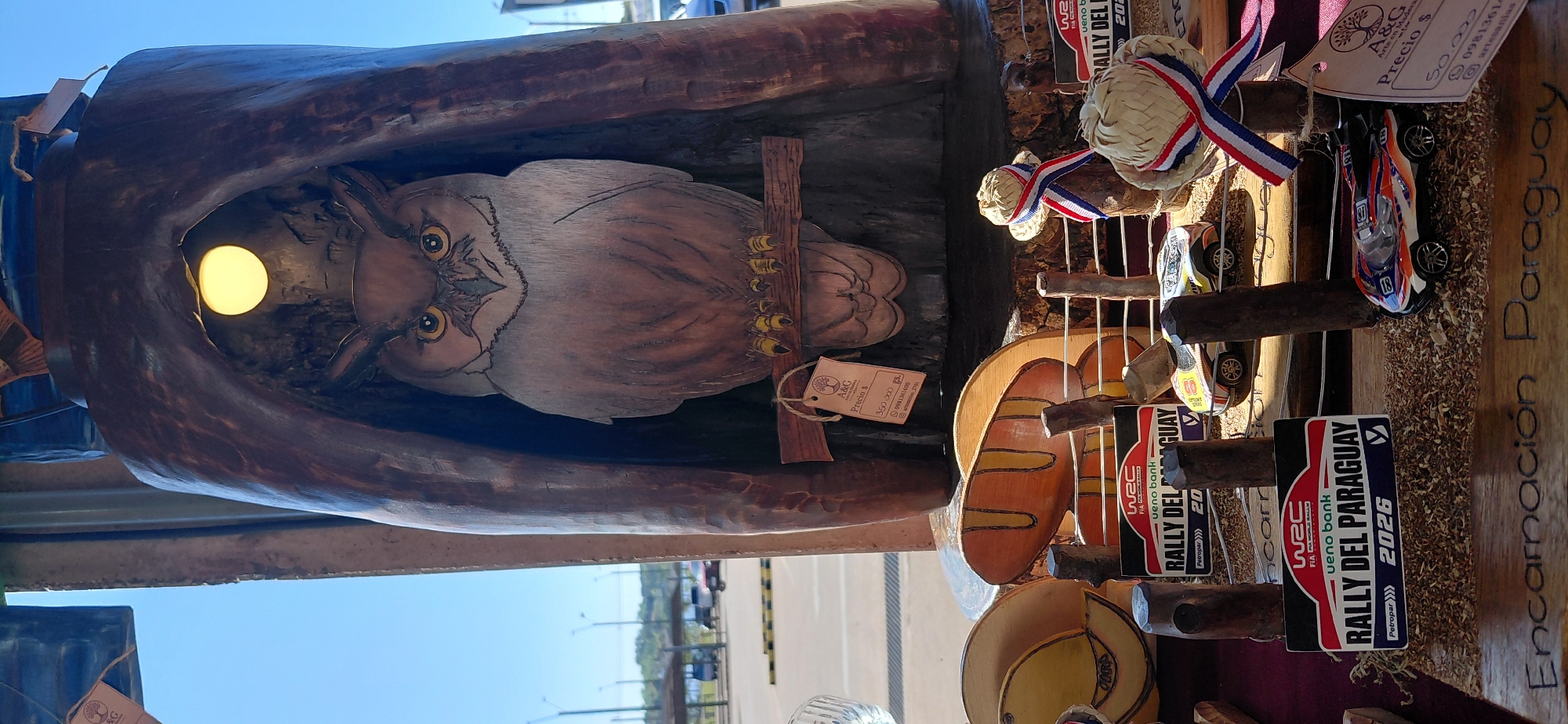 The image shows a wooden sculpture of an owl perched on a branch inside a hollowed-out log. The sculpture is lit by a small yellow light bulb within the log. Below the owl, there is a price tag with "A&G Arte en Madera" and "Precio $" written on it. Further down, there are several miniature toy cars and decorative elements, including small straw hats adorned with red, white, and blue ribbons, and signs for "WRC Rally del Paraguay". A wooden sign at the bottom of the image reads "Encarnación Paraguay". The background suggests an outdoor setting with a parking lot and some vehicles visible. The scene appears to be a stall or display at a market or craft fair in Encarnación, Paraguay.