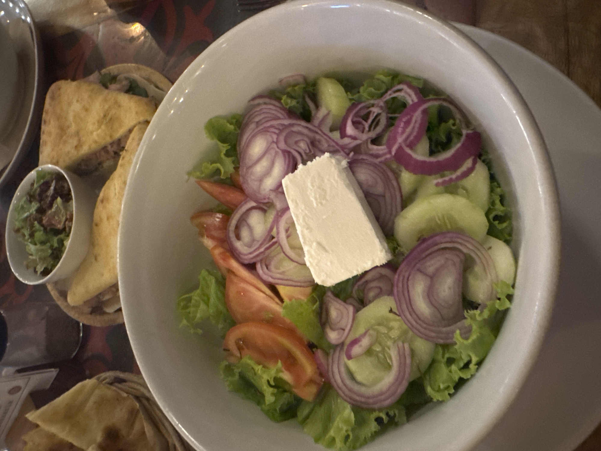 The image presents a dining table scene, focusing on a white bowl filled with a salad composed of lettuce, sliced cucumbers, tomato wedges, and prominent red onion rings. At the center of the salad is a rectangular block of white cheese, presumably feta. Adjacent to the bowl, there are portions of pita bread and a small bowl containing what appears to be a dip. The setting seems to be a restaurant or indoor dining area, given the typical arrangement of dishes and the presence of a table. No people are directly visible in the frame. Specific location in the city of Kuta Selatan, Indonesia cannot be determined without further information.
