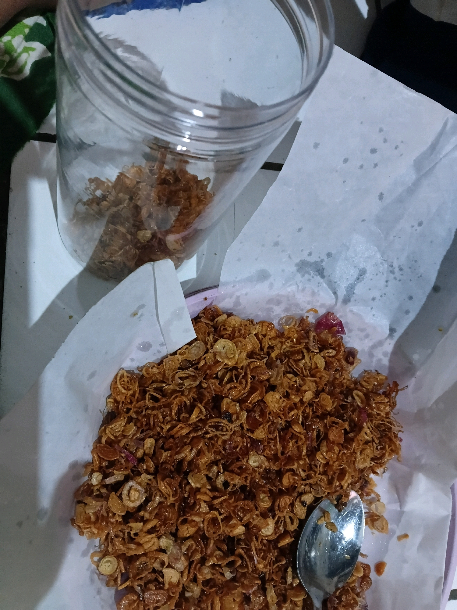 A close-up, high-angle shot shows a pile of fried shallots and garlic on white paper, with a glass jar containing more of the fried mixture in the background. A silver spoon rests on the pile of food. The food is golden brown and appears crispy. The setting is likely a home kitchen or dining area in Wadungasri, Indonesia, given the context. The lighting is natural and diffused, creating soft shadows. The overall impression is one of homemade food preparation or serving.
